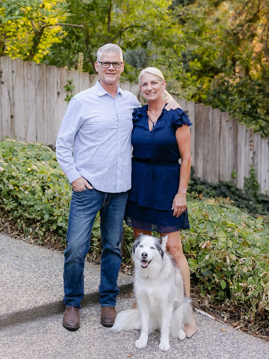 A couple standing outdoors with their Siberian Husky dog, in a garden with greenery and a wooden fence, on a paved pathway.