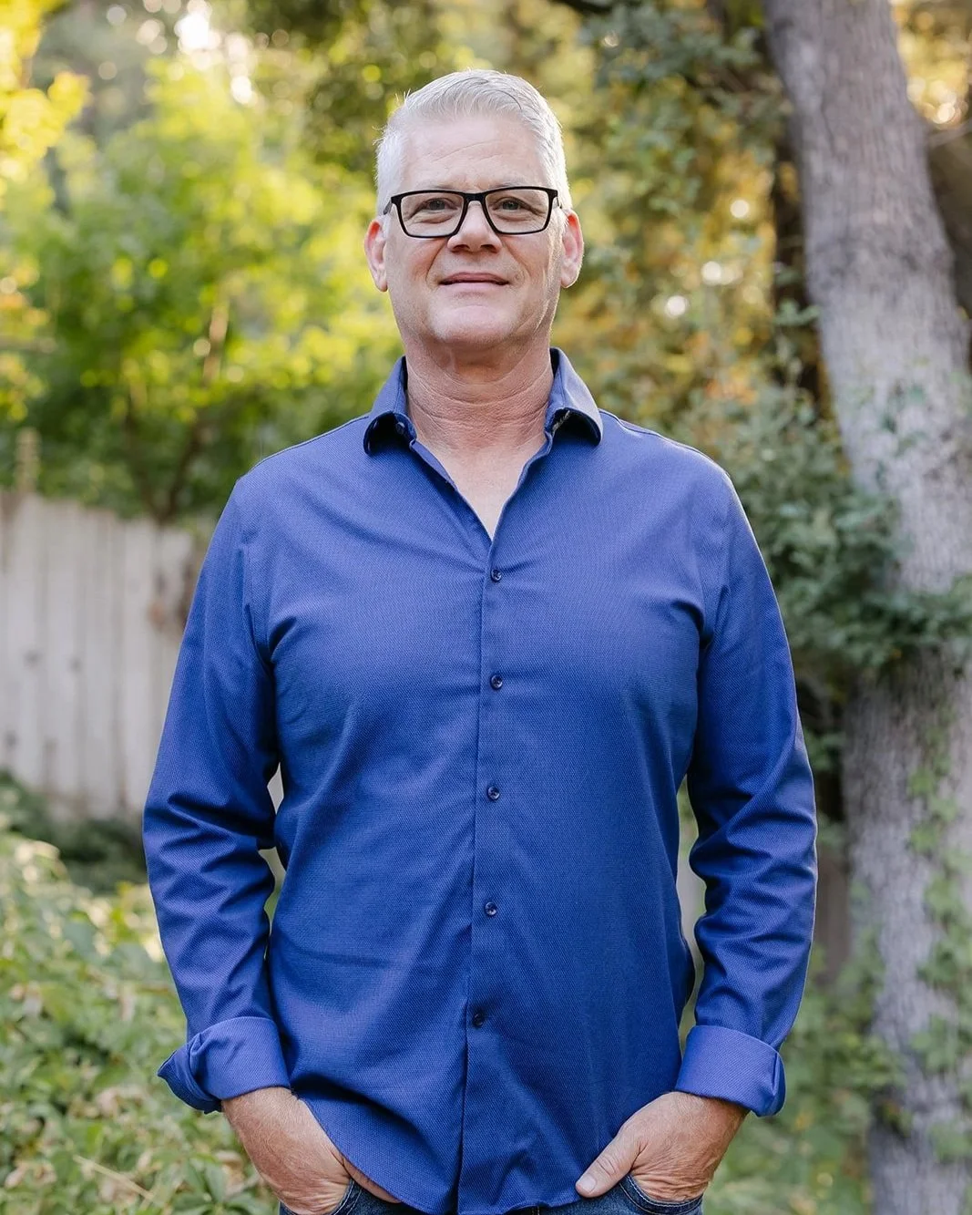 A man with glasses and short gray hair stands outdoors in a garden or park with green trees and a wooden fence in the background. He is wearing a blue button-up shirt with rolled-up sleeves and has his hands in his pockets, smiling at the camera.