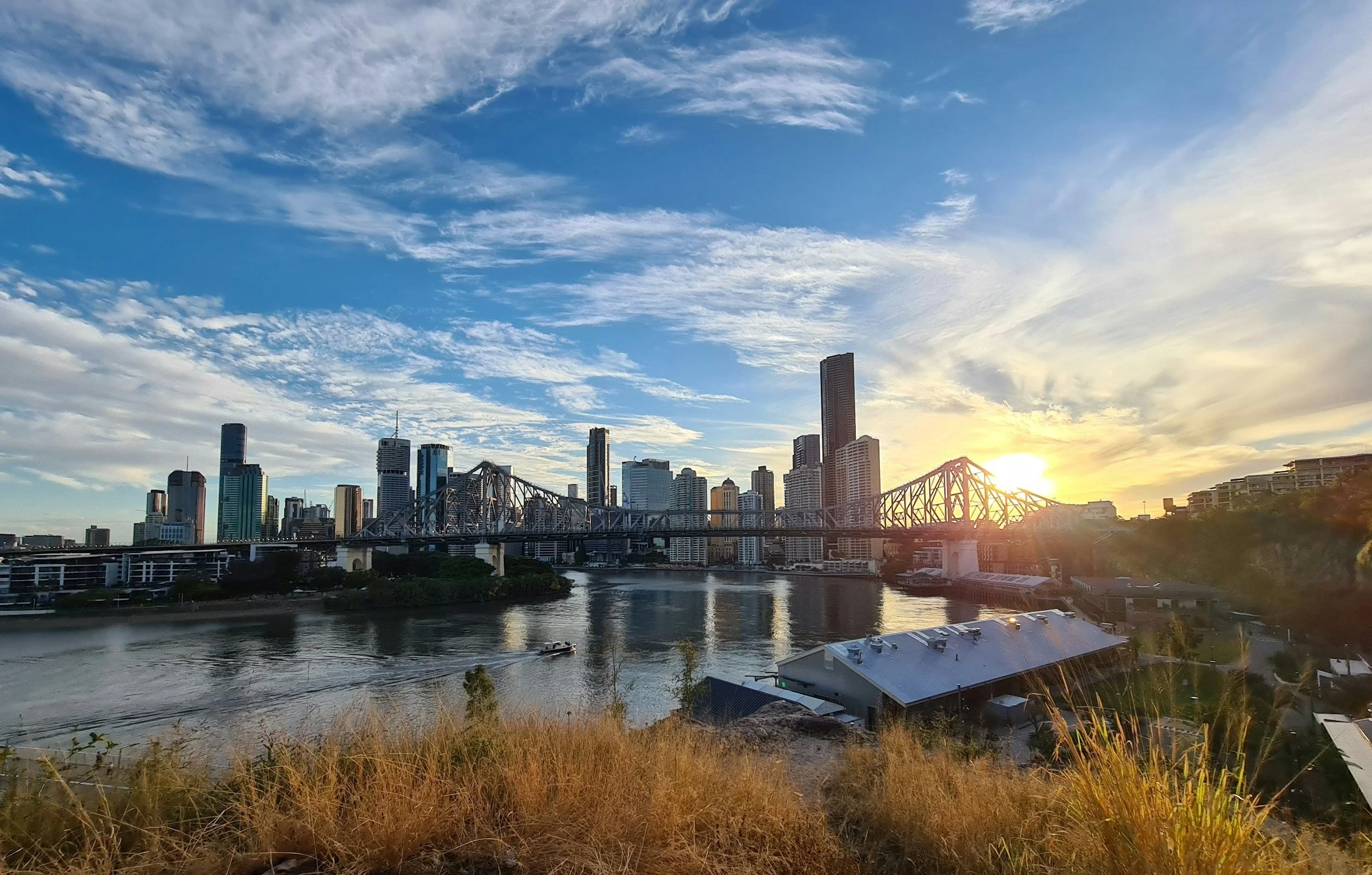 City skyline at sunset with tall buildings, a river, and a bridge, under a partly cloudy sky.