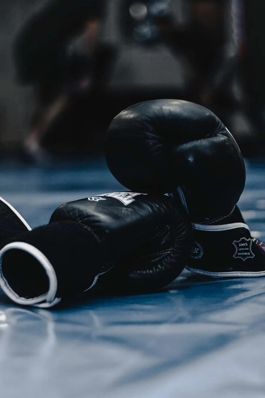 Boxing gloves and headgear resting on a gym floor with a martial arts training session in the blurred background.
