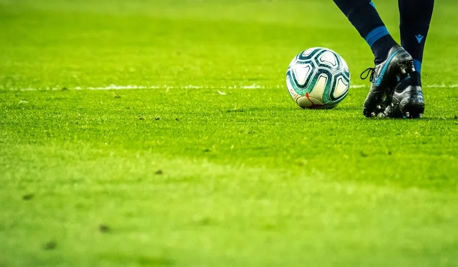 Close-up of a soccer player’s feet with black cleats on a green field, about to kick a white soccer ball with teal and black designs.