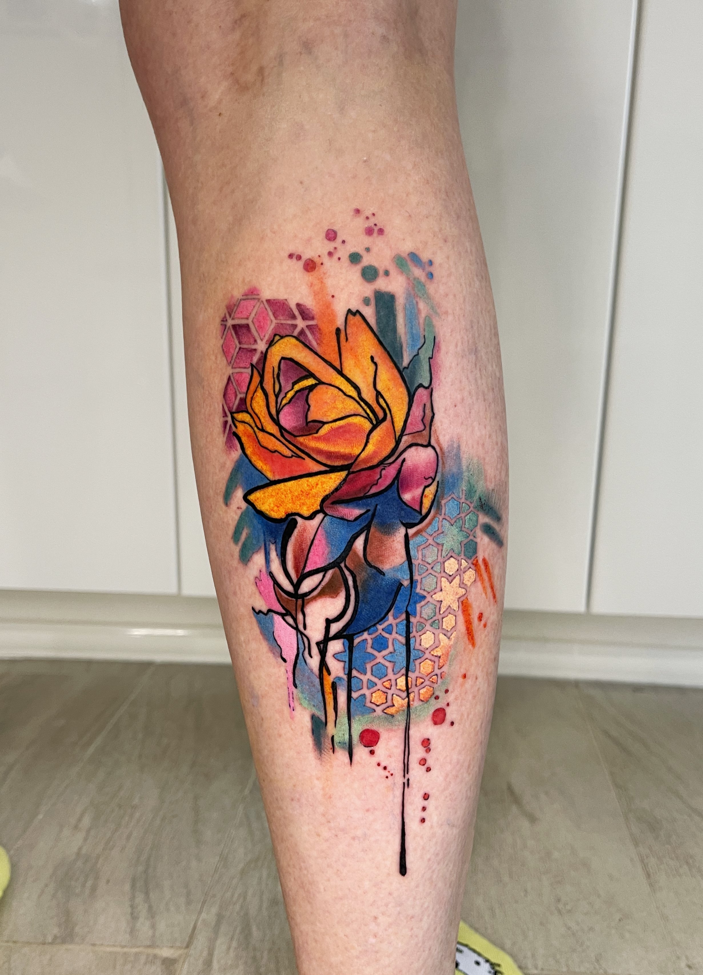 Colorful tattoo of a rose with abstract watercolor background and geometric patterns on a person's thigh