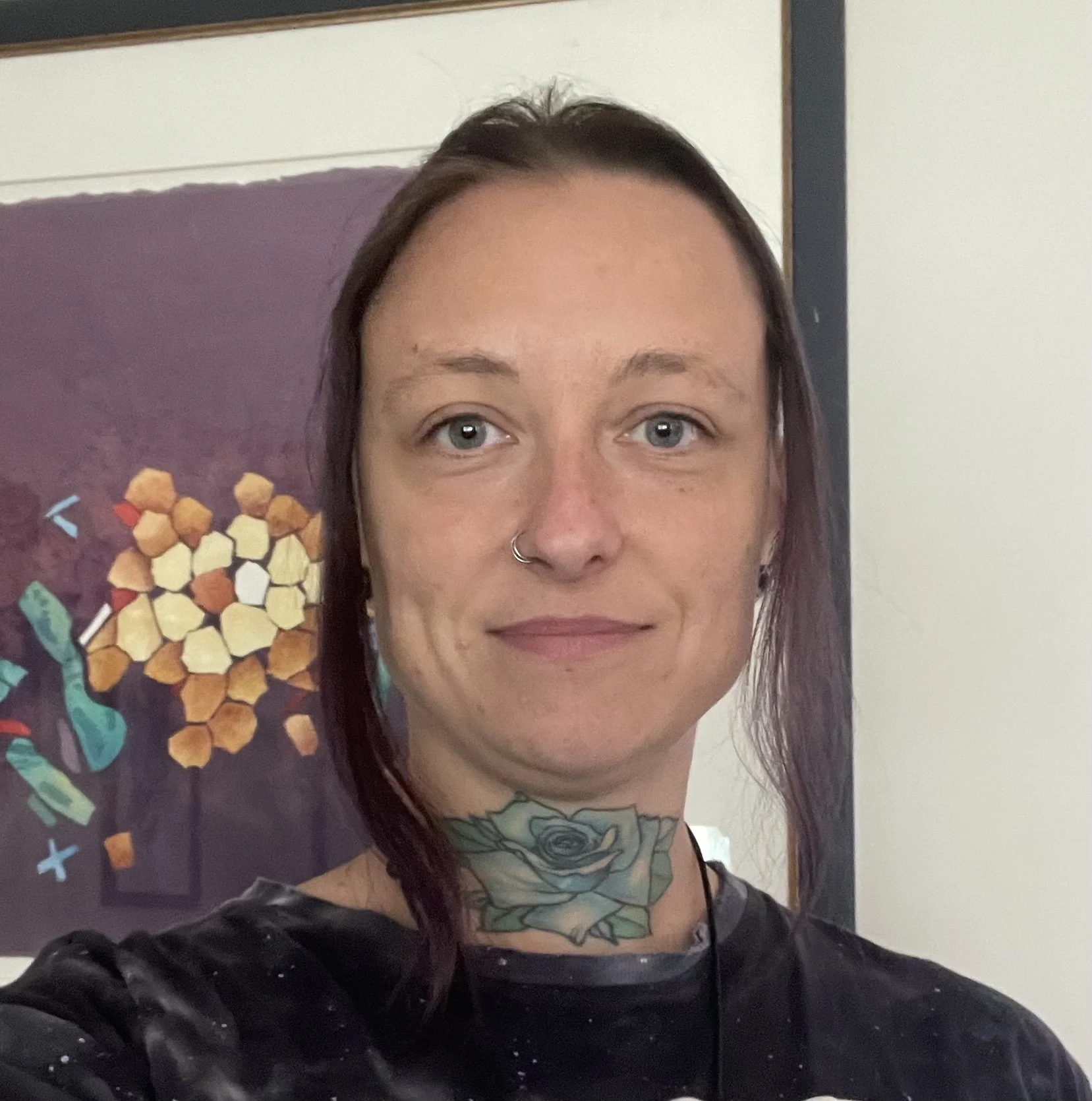 A woman with dyed purple hair, a nose ring, and a large floral tattoo on her neck, taking a selfie in front of a framed artwork featuring abstract shapes and colors.