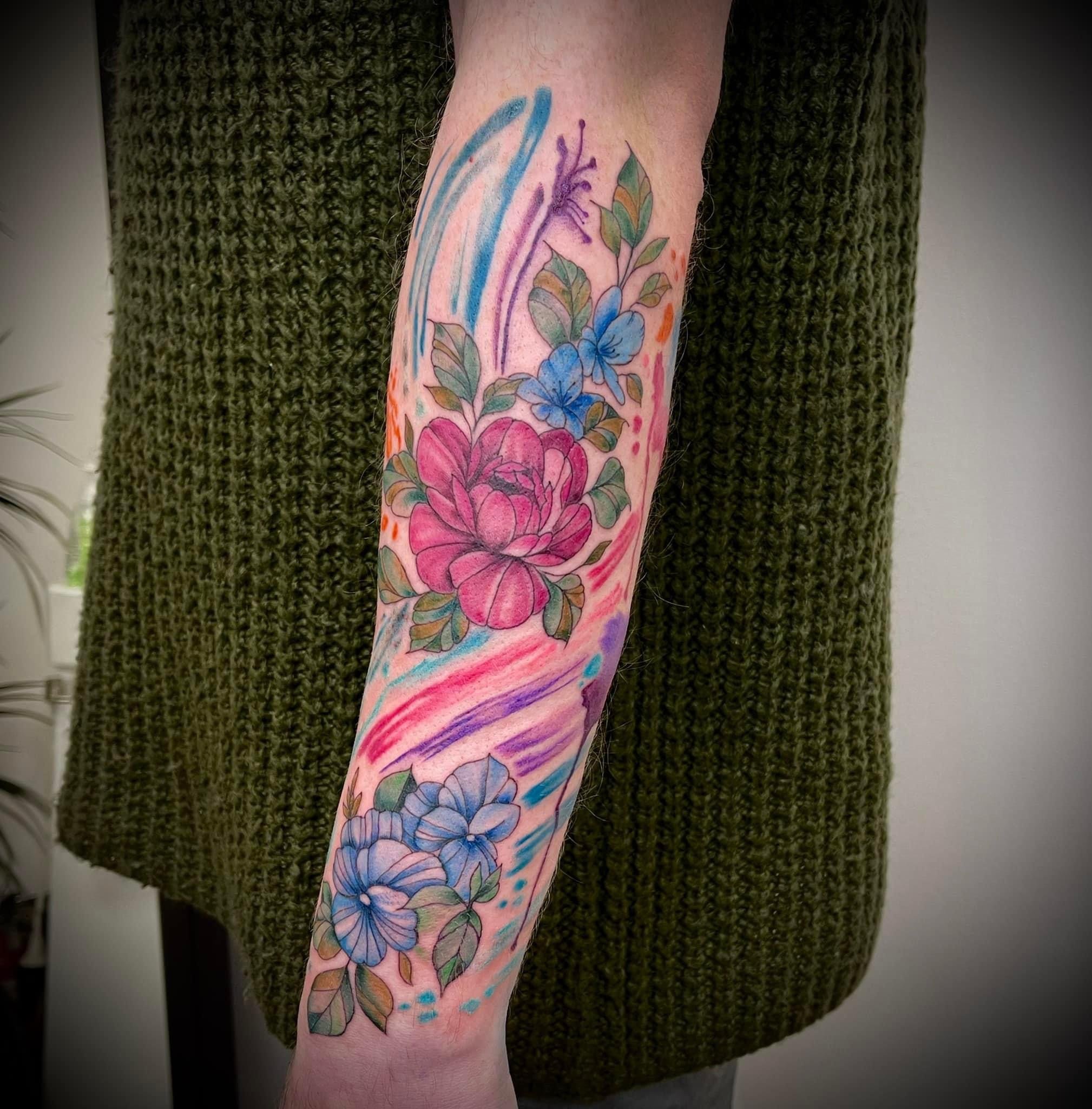 Colorful watercolor floral tattoo on person's forearm with blue, pink, purple, orange, green hues, and leafy accents.