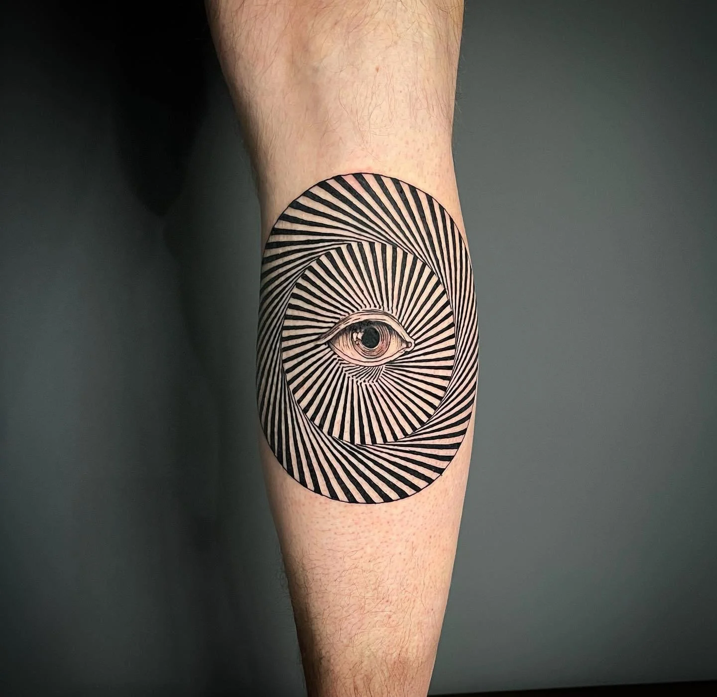 Tattoo of a detailed eye at the center of a circular pattern with black and white radial lines on a person's leg.