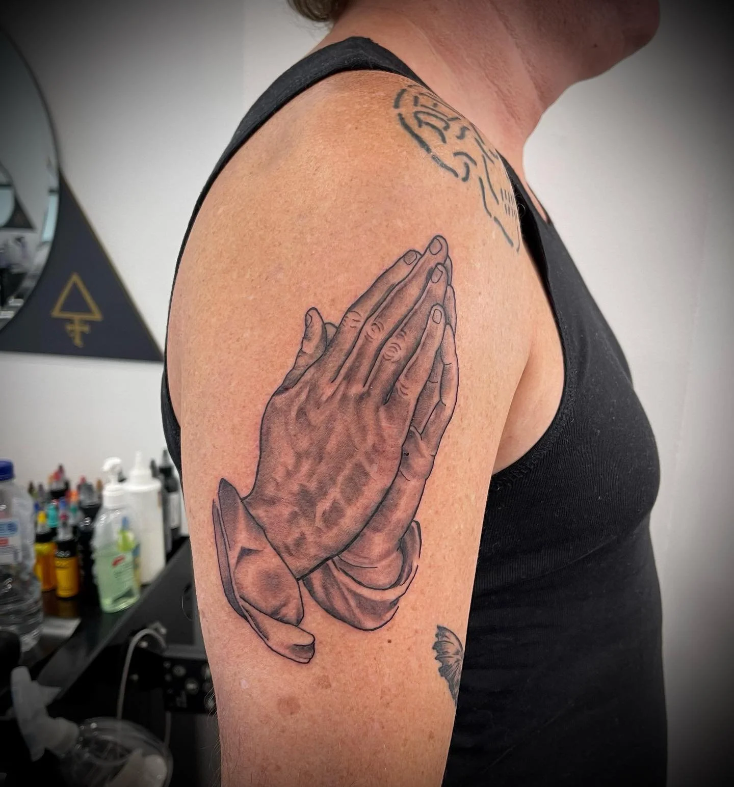 Tattoo of two praying hands on a person's upper arm and a tattoo of a head on the shoulder, in a tattoo parlor with tattoo supplies in the background.