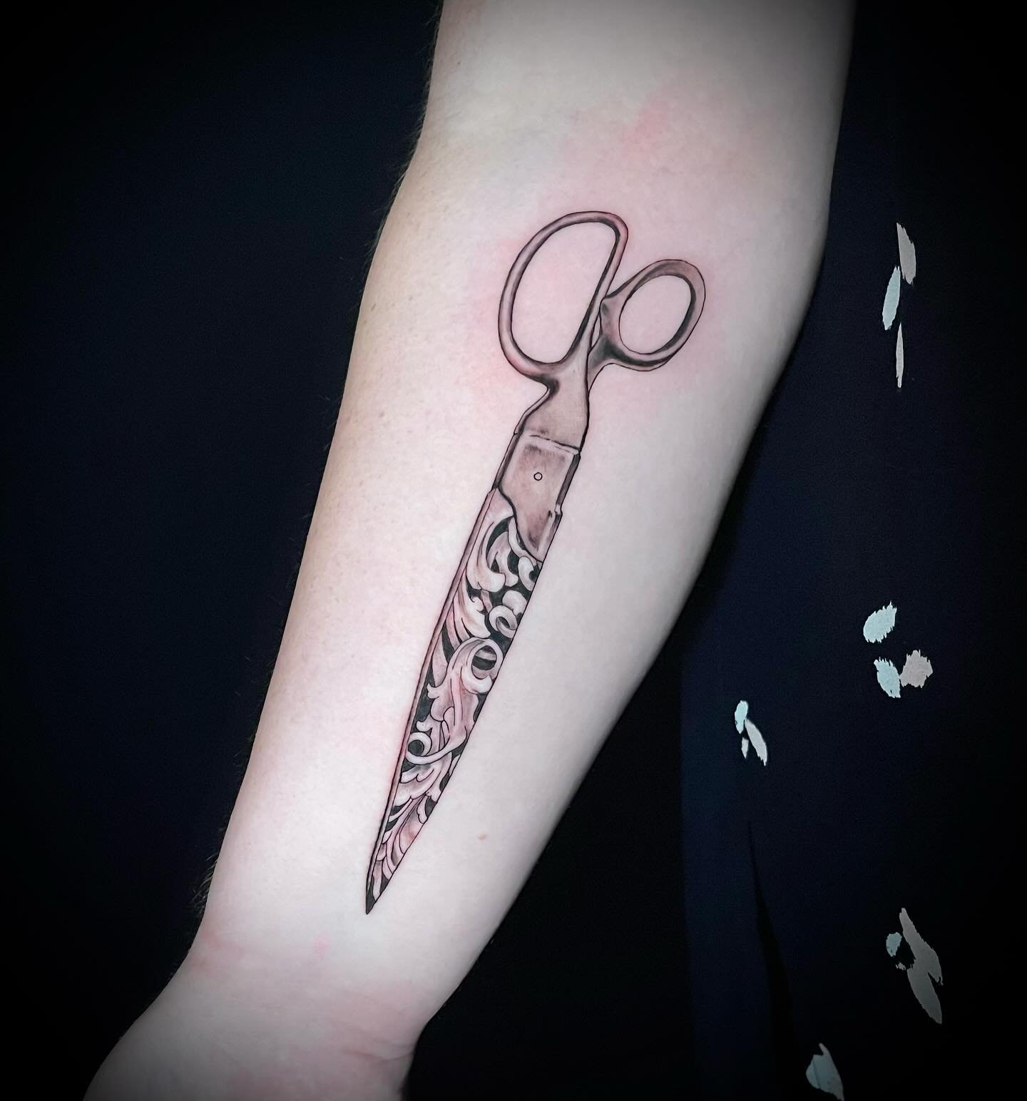 Tattoo of scissors above a detailed, swirling pattern on inner forearm.