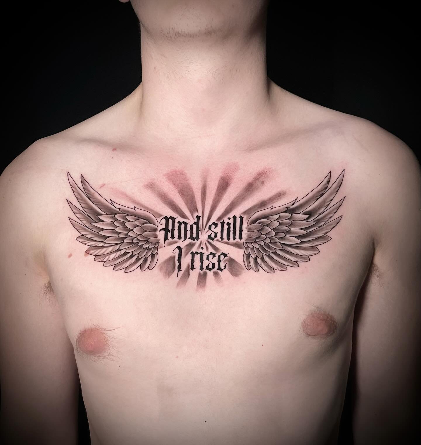 A tattoo on a person's chest featuring angel wings with shading and the phrase 'And still I rise' in Gothic font.