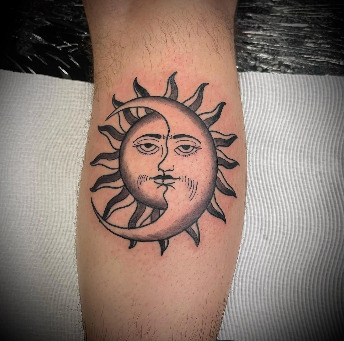 Tattoo of a sun with a face on a person's arm. The sun has a moon shape on the left side, and the face has one eye open and one eye closed.