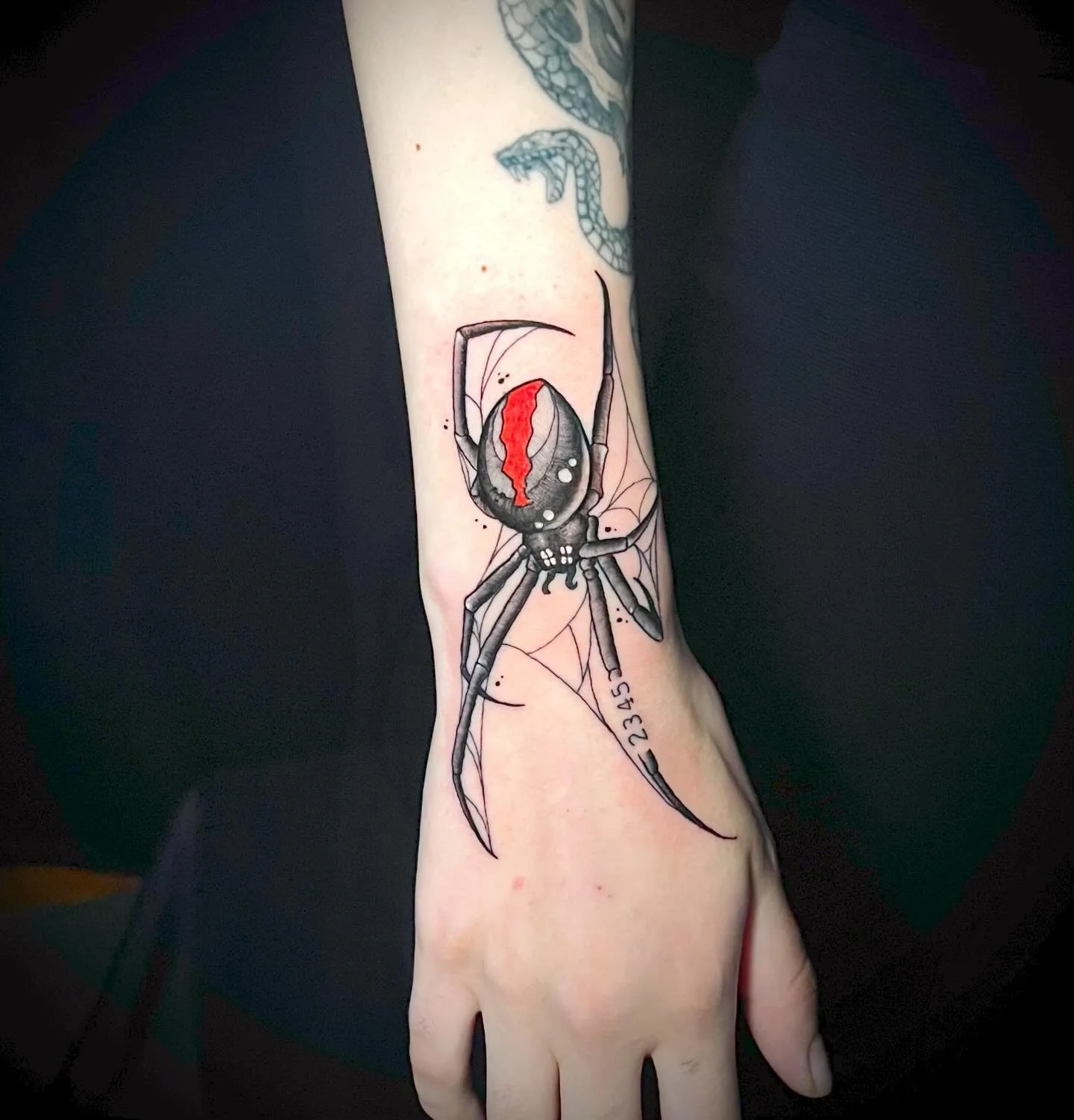 A tattoo of a black widow spider with a red hourglass on its abdomen, depicted on a person's forearm. The spider's body is detailed with black and gray shading, and it appears to be sitting on a web.