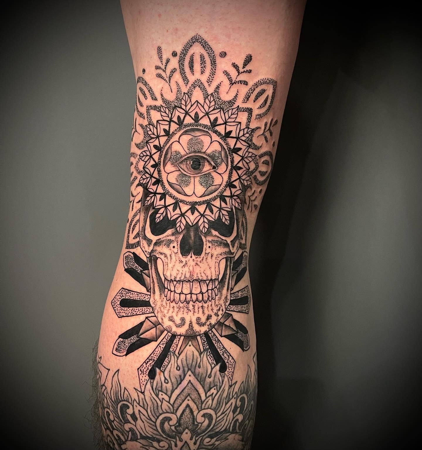 Black and gray tattoo of a human skull with detailed line work, floral motifs, a central eye, and geometric patterns on the person's arm.