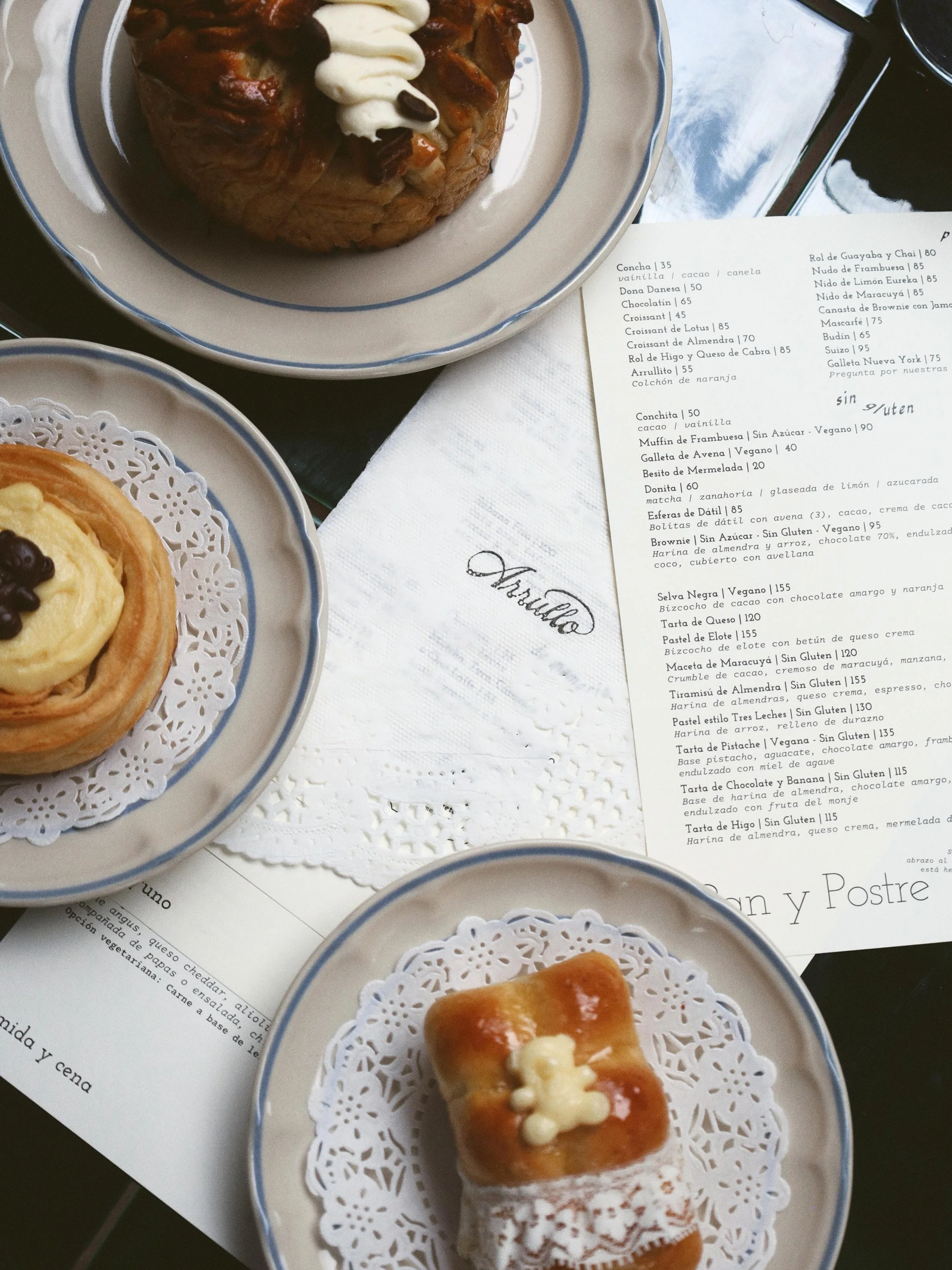 Varias porciones de postres regulares, veganos y sin gluten en platos decorados con adornos de papel, incluyendo pasteles con glaseado, crema y frutas, sobre el menú de Arrullo.