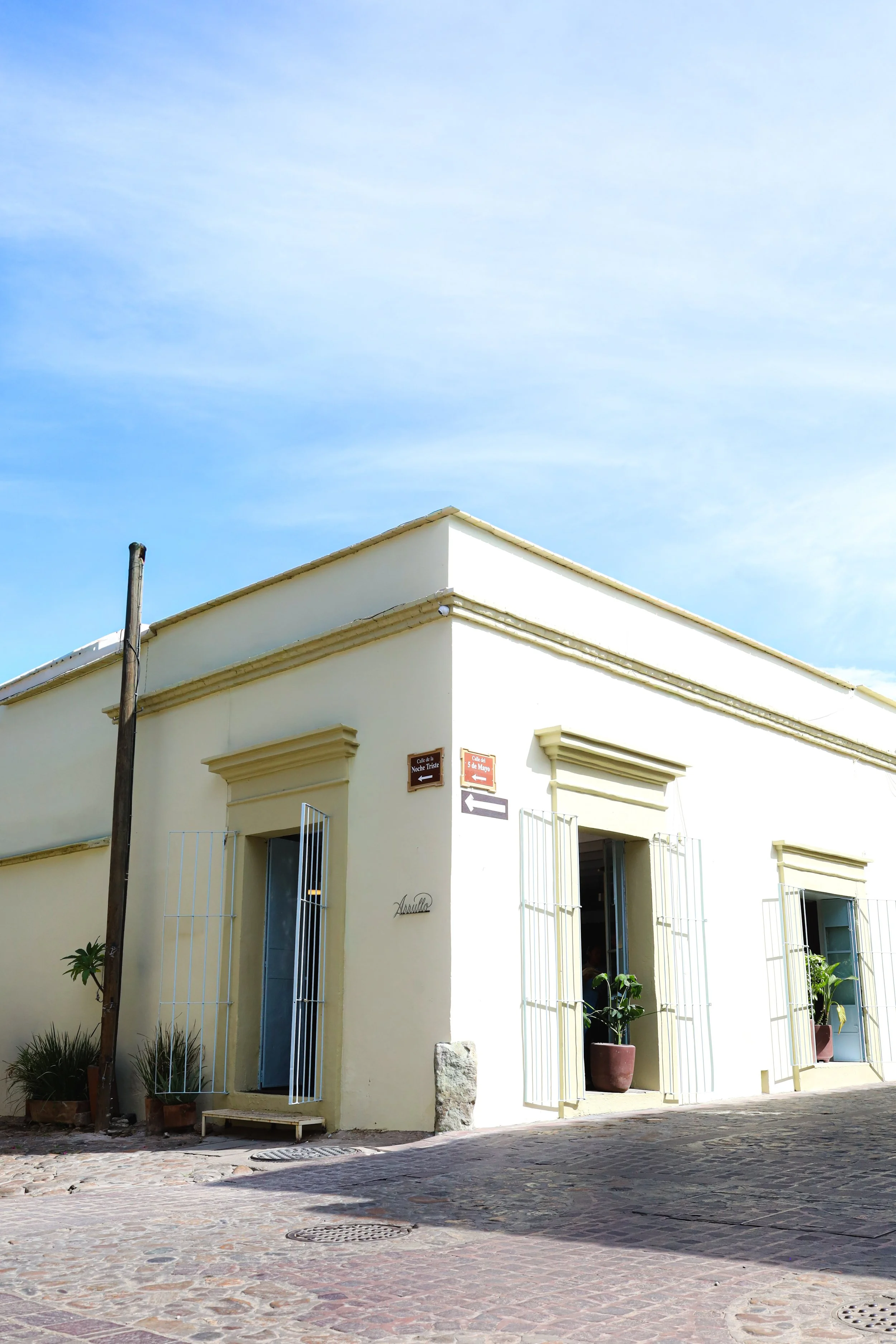 La casa de Arrullo. Un edificio de color crema con ventanas abiertas y plantas en macetas, en una calle empedrada llamada 5 de Mayo en el barrio de Jalatlaco, Oaxaca con el cielo azul de fondo.