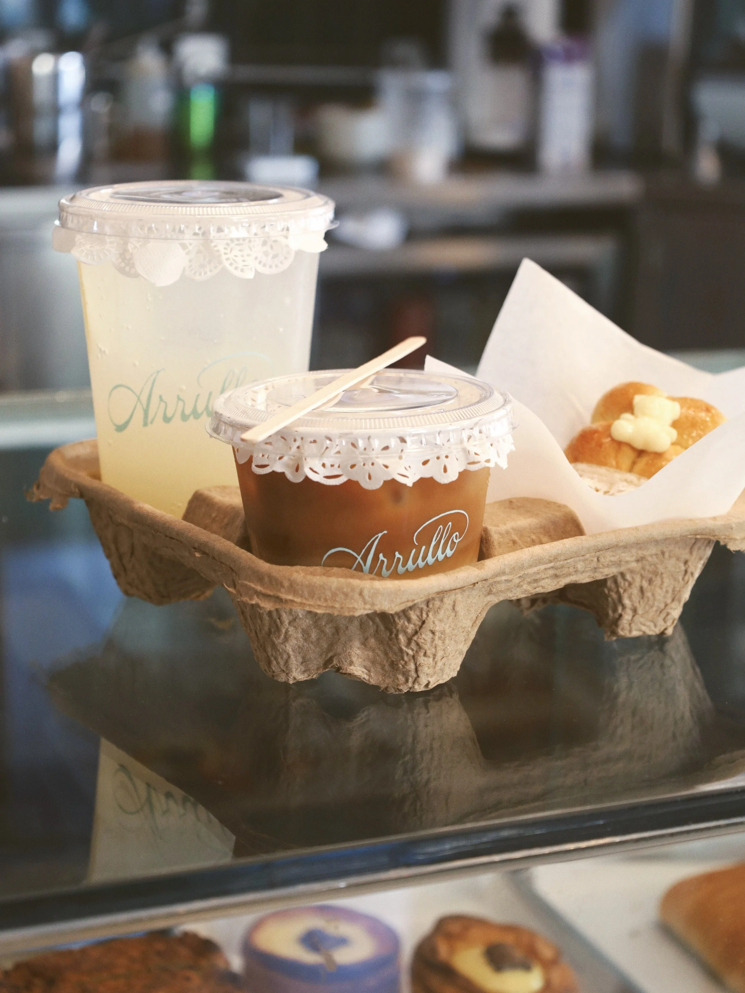 Varios envases transparentes con tapa para recoger en tienda o para llevar, uno con bebida y otro con postre, en una bandeja de carton, sobre una vitrina de nuestra cafetería y panadería en Oaxaca.