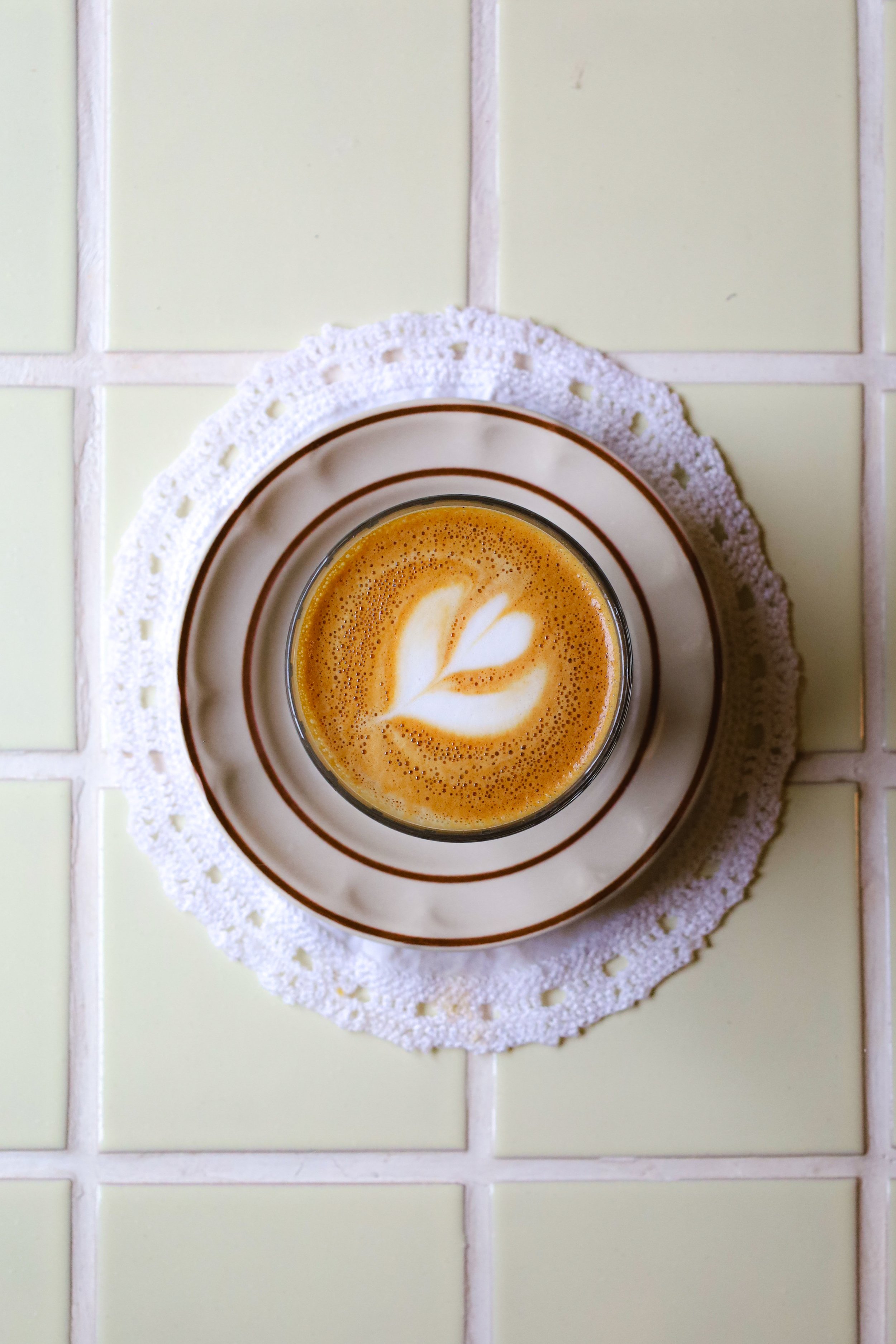 Taza de café con leche con arte latte en la espuma de leche, sobre un platillo con borde de rayas marrones, en un mantel de encaje blanco sobre una mesa con baldosas de color beige claro. Opciones de café en Arrullo Oaxaca.