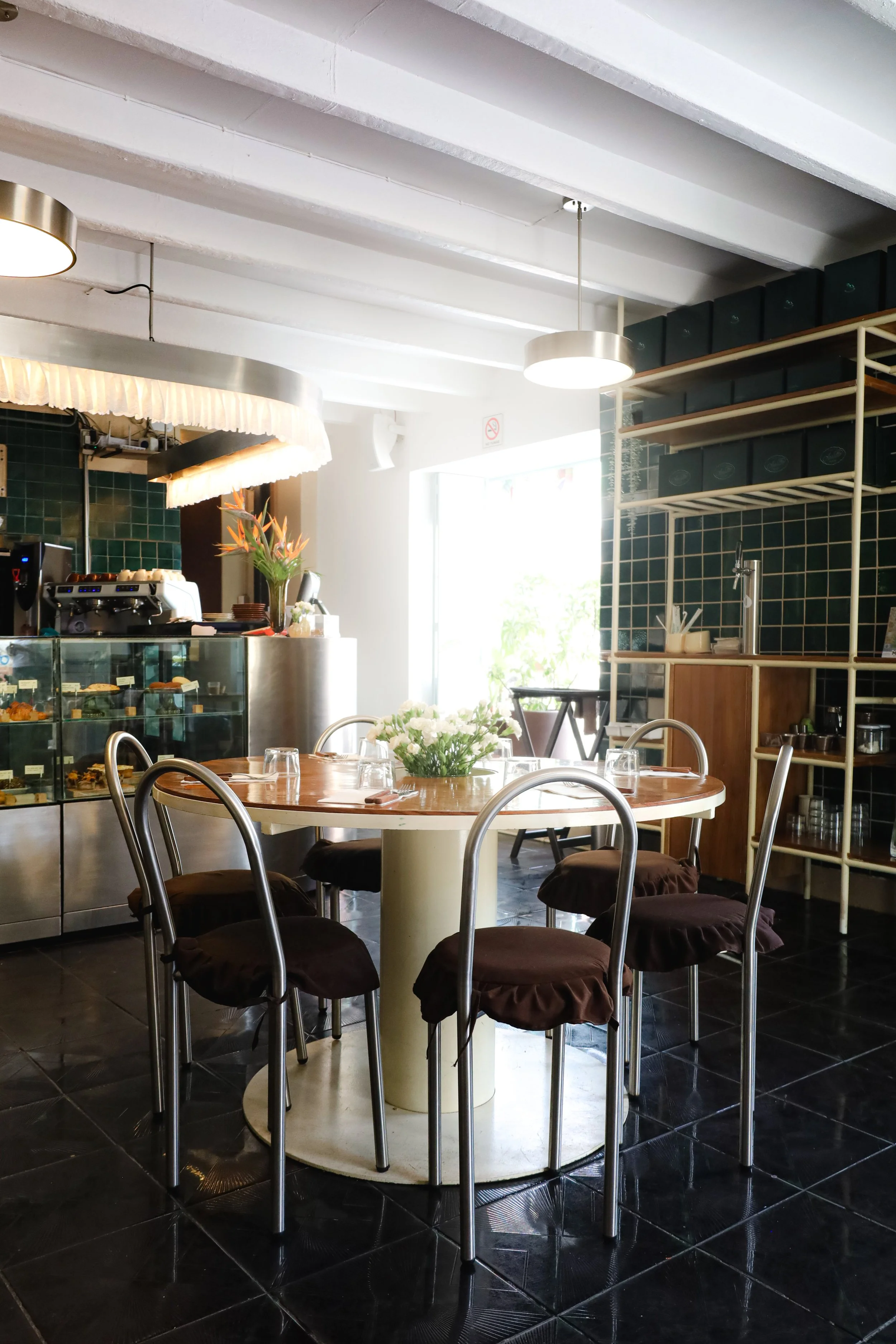 Interior de Arrullo Oaxaca con una mesa redonda de madera, sillas con cojines oscuros, un mostrador con una vitrina de panes y postres, plantas con flores y una repisa con cajas en la pared, luz natural entrando por una ventana