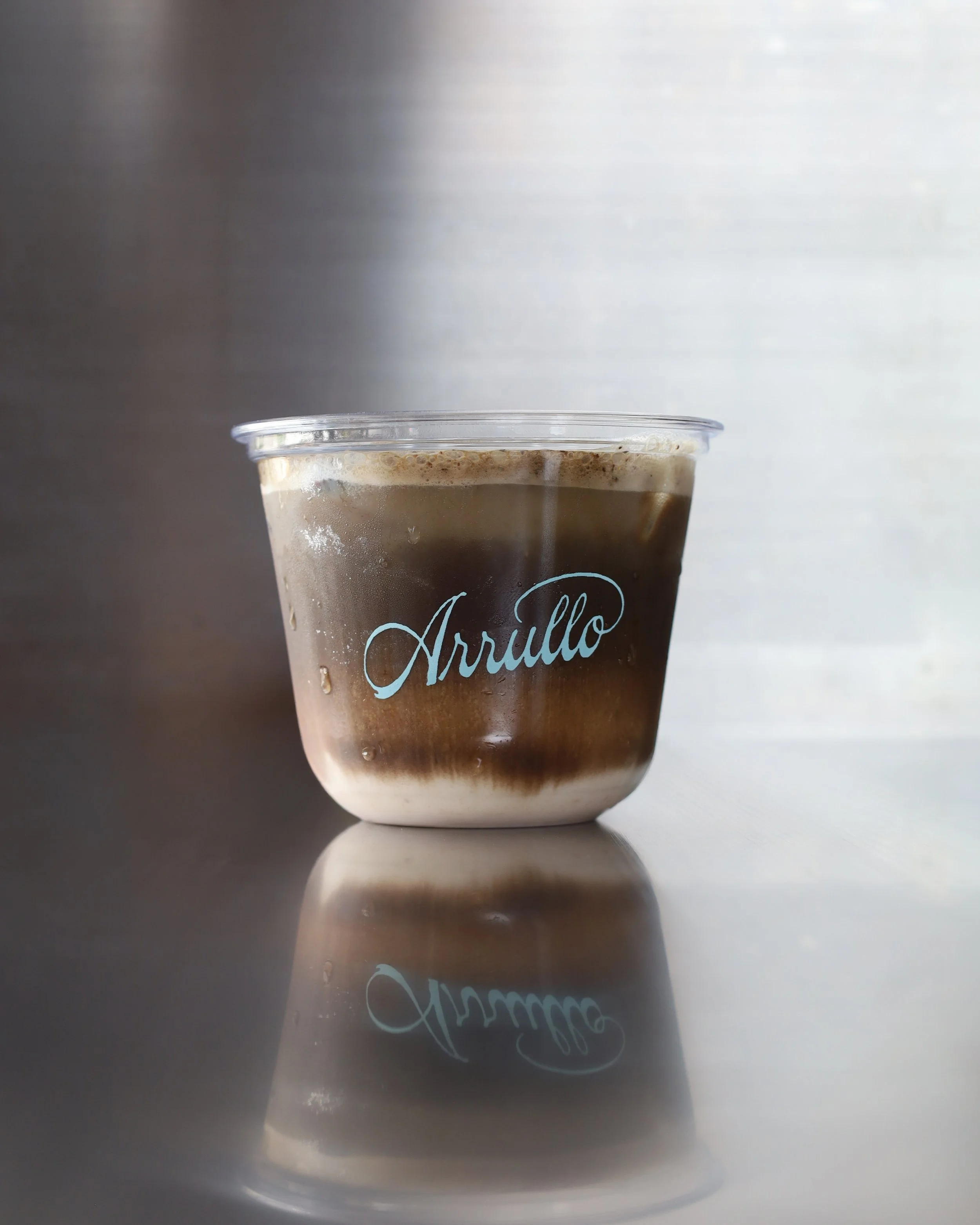Vaso de bebida fría lleno de horchata de la casa y una carga de te hojicha, con la palabra 'Arrullo' escrita en letras cursivas en el vaso.