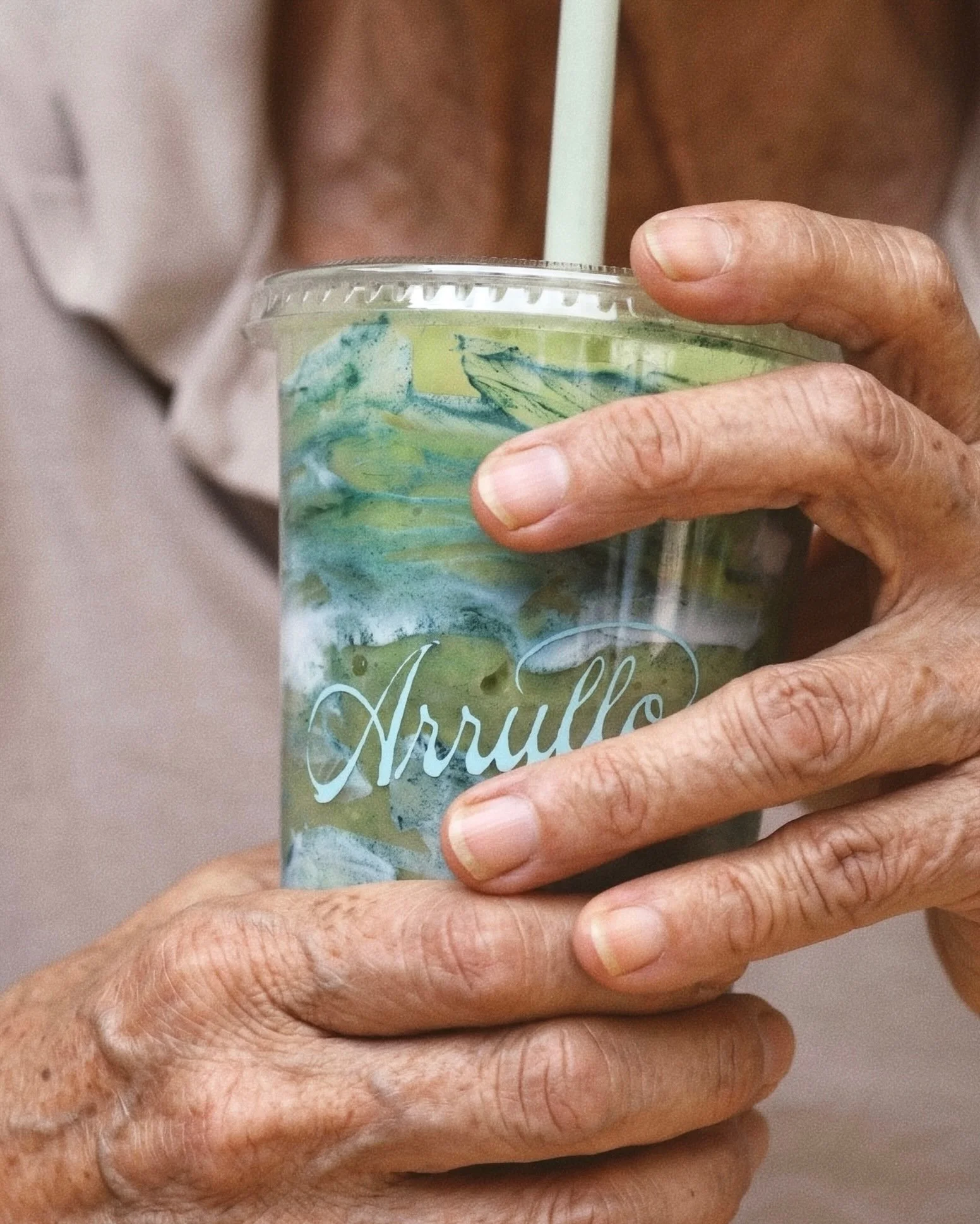 Mano de persona mayor sosteniendo un vaso de plástico lleno de uno de nuestros batidos de lacasa con decoraciones de color verde y azul, con la palabra 'Arrullo' en el vaso.