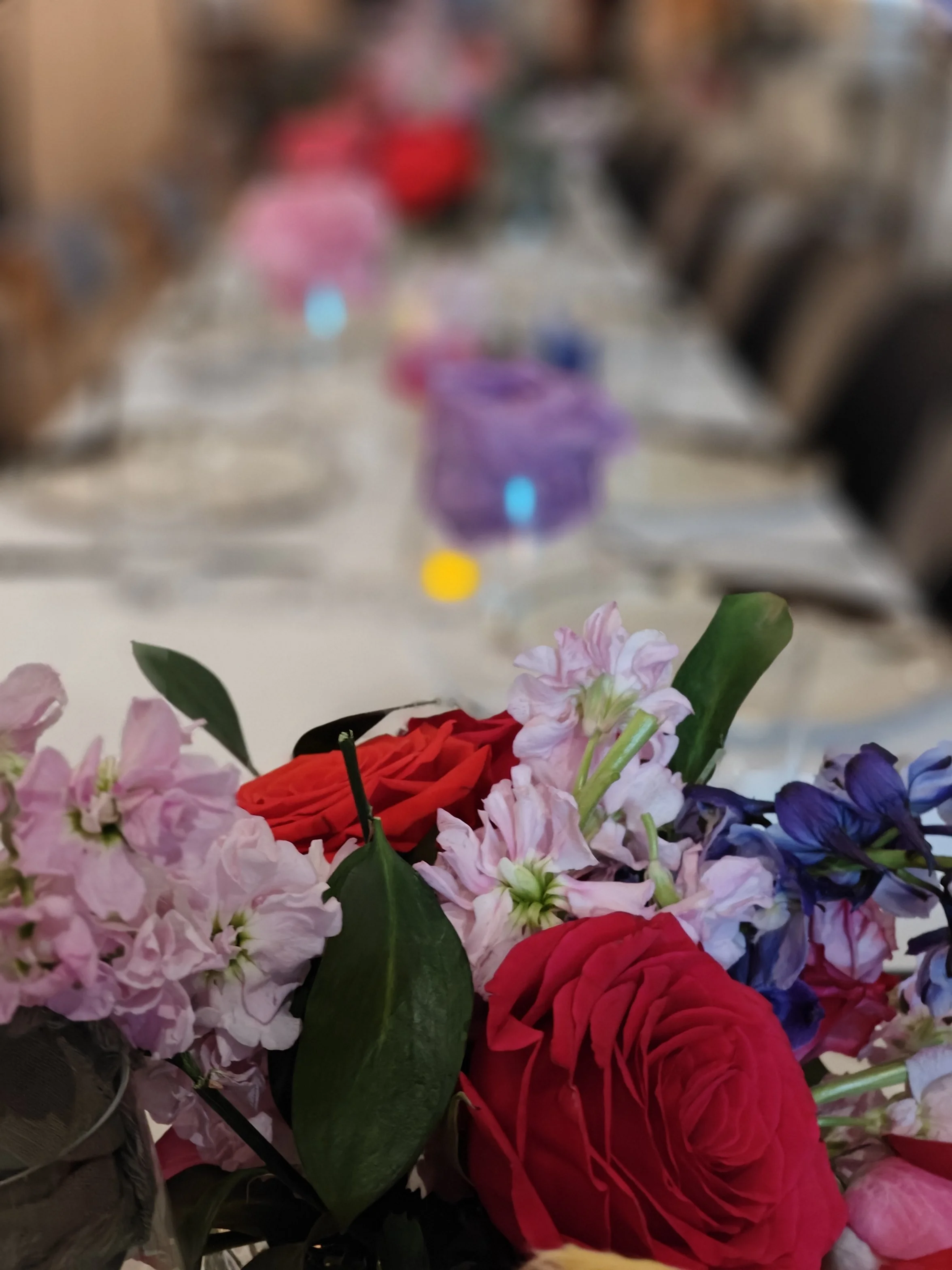Close-up of a bouquet of pink, red, purple, and white flowers with green leaves in the foreground, blurry background.