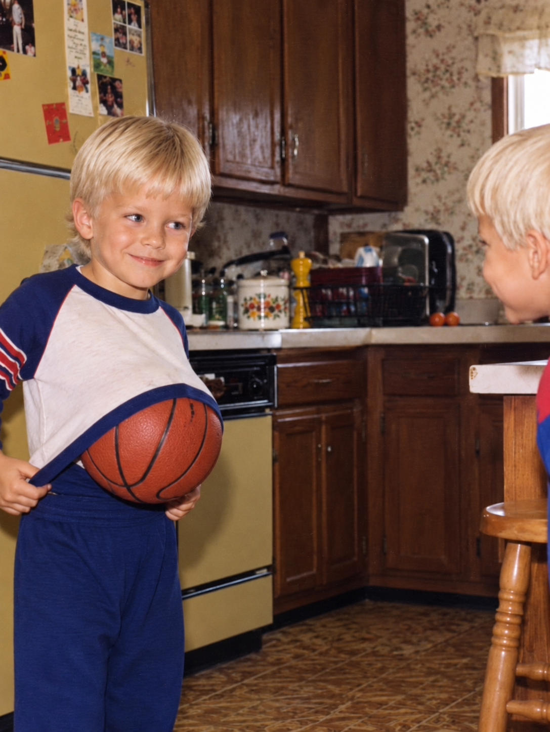 A childhood photo of William smiling with a basketball tucked under his shirt in the family kitchen.