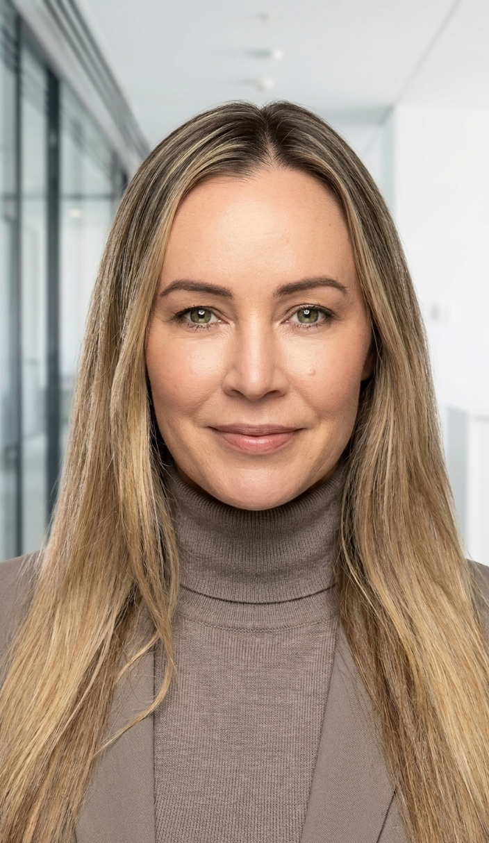 A professional woman with long blonde hair, wearing a tan turtleneck and blazer, smiling in an office corridor with glass walls.