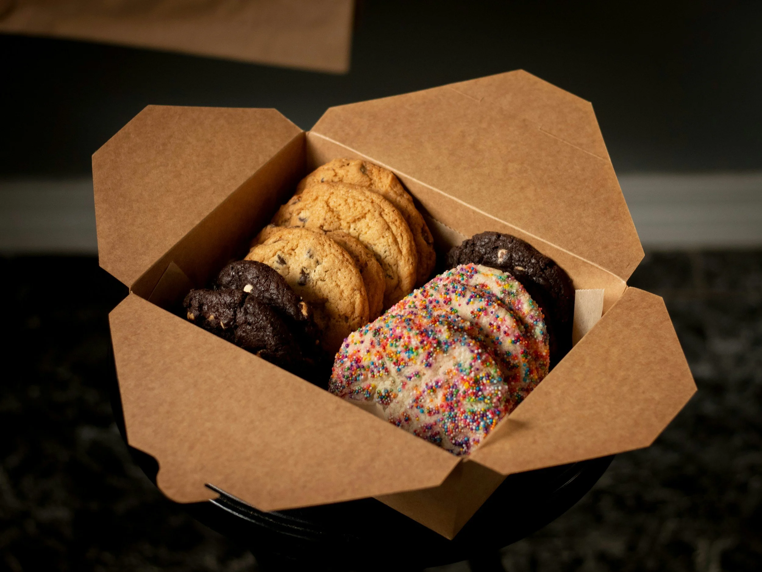 A cardboard box filled with assorted cookies, including chocolate chip, sprinkled, and double chocolate cookies.