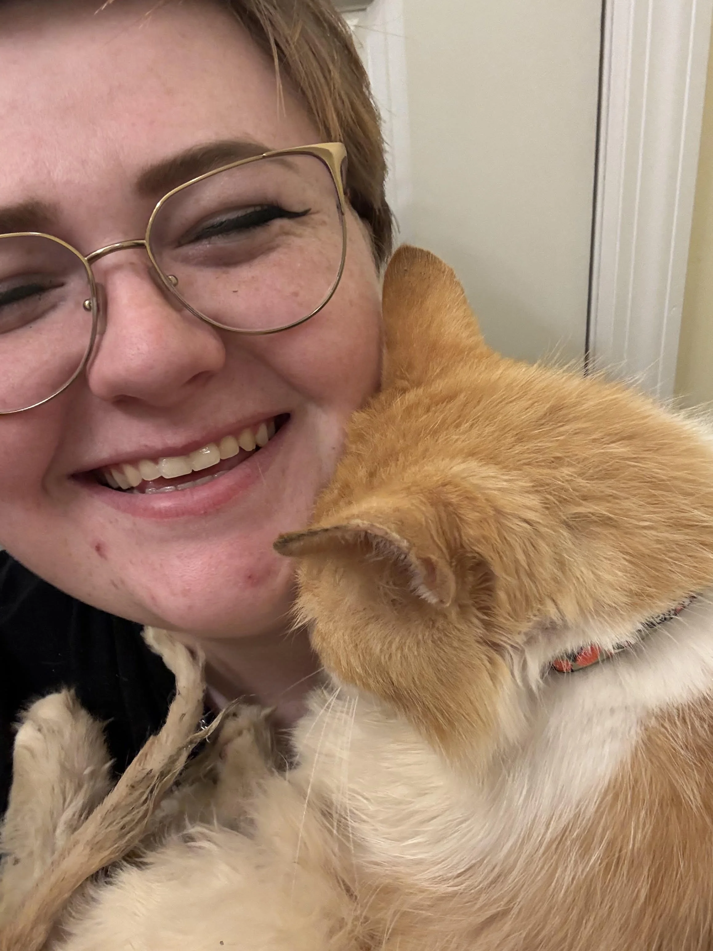 Close-up of a smiling woman with glasses and an orange tabby cat sitting on her shoulder.