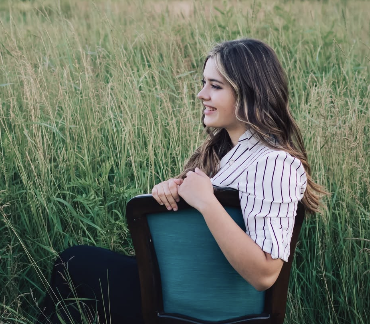 A young woman with long wavy hair, wearing a white top with purple stripes, sitting in a field of tall grass and holding a dark wooden chair with a teal cushion, smiling and looking to her left.