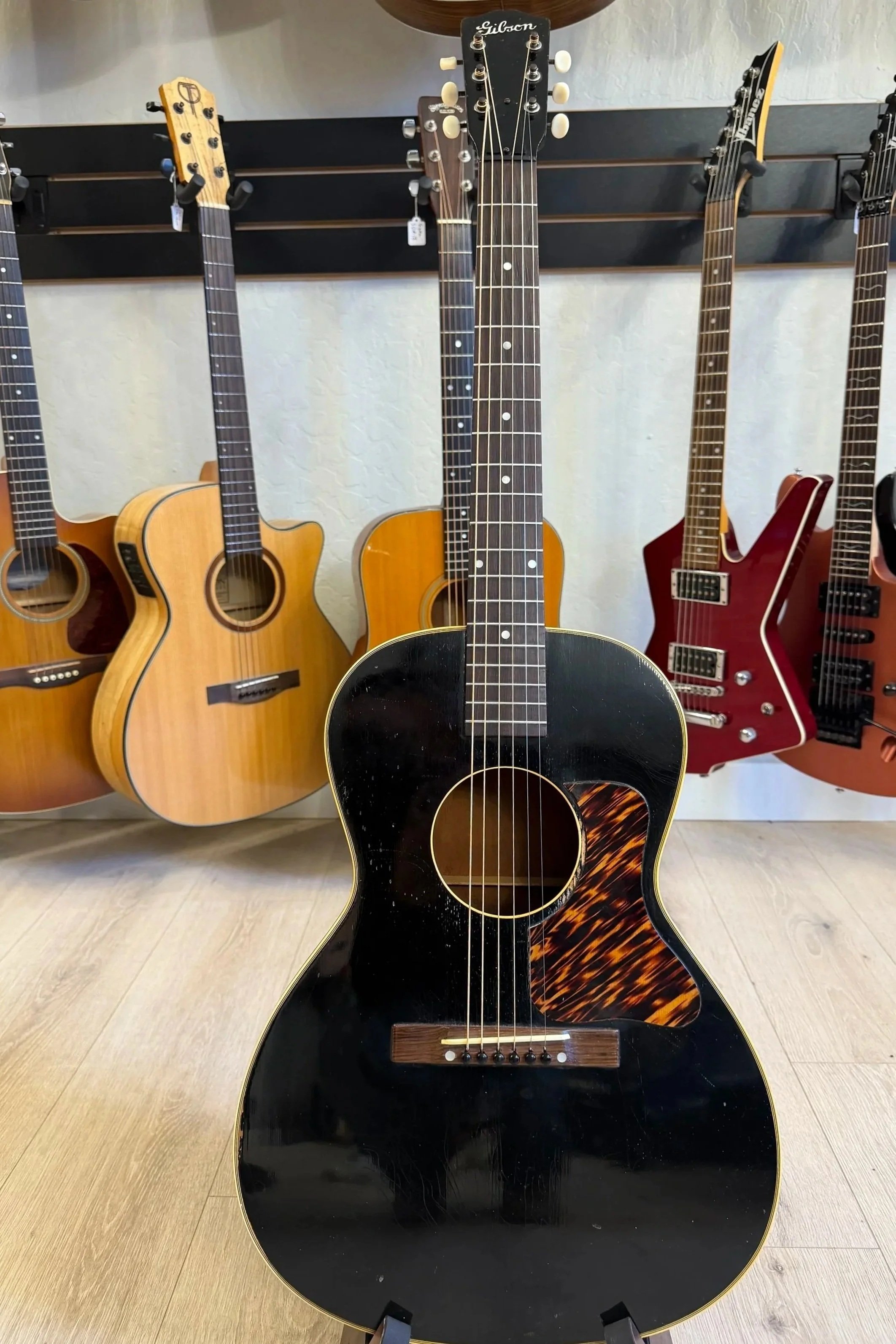 A display of various guitars on a wall, with a central black acoustic guitar in the foreground featuring a tortoiseshell pickguard.