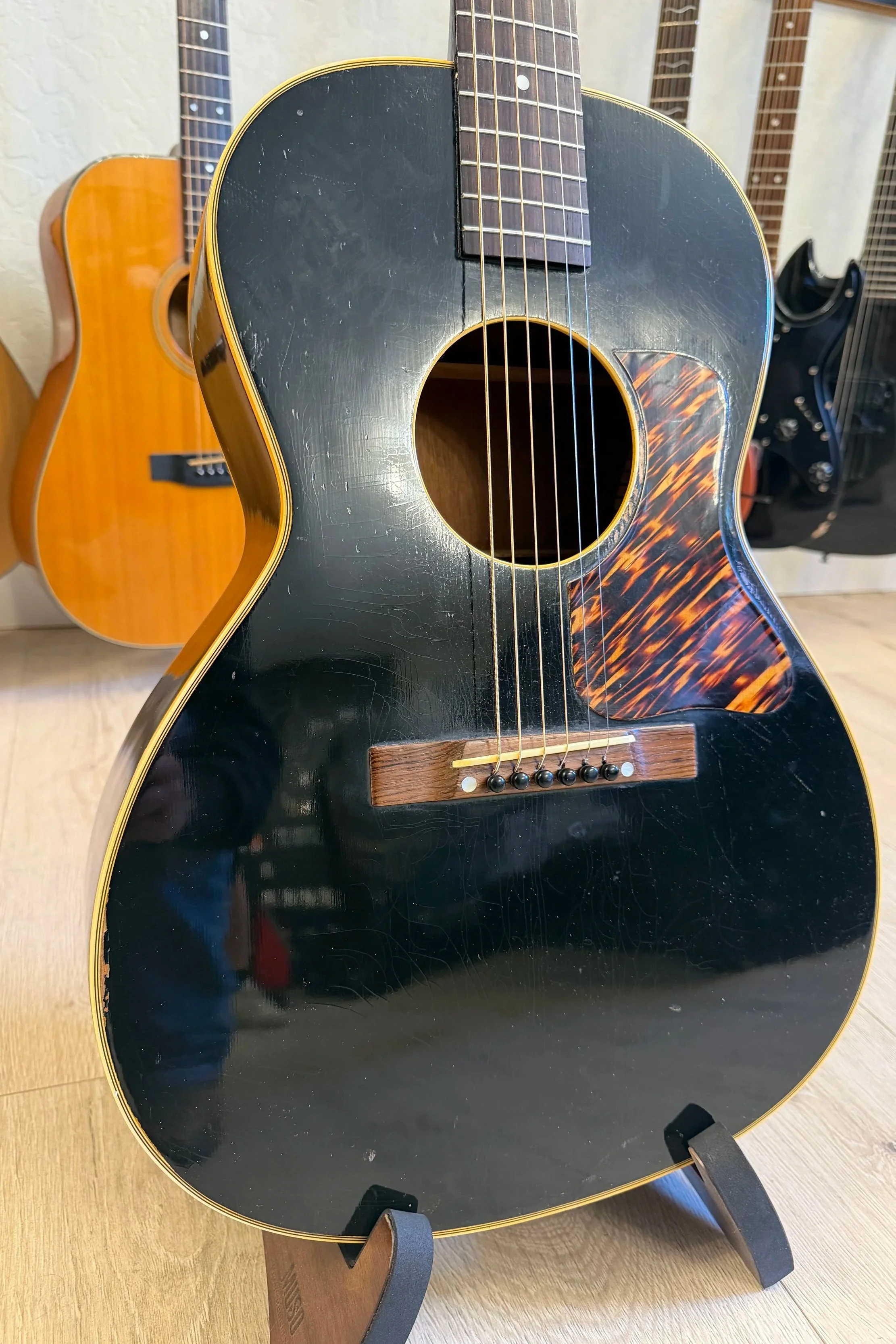 Black acoustic guitar with a tortoise shell pickguard and some scratches, displayed on a black stand on a wooden floor, with lighter-colored guitars visible in the background.