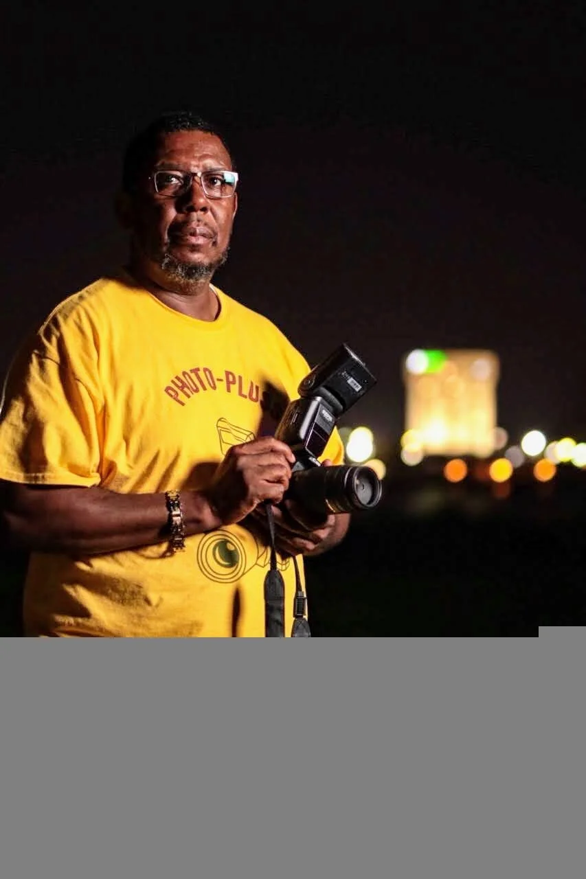 A man wearing glasses and a yellow T-shirt holding a camera at night with a blurred city skyline in the background.