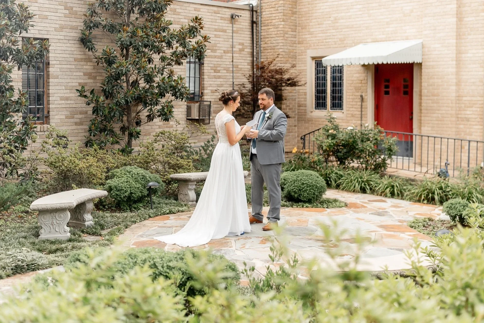 A bride and groom exchange vows in a garden courtyard with brick walls, green bushes, and stone benches.