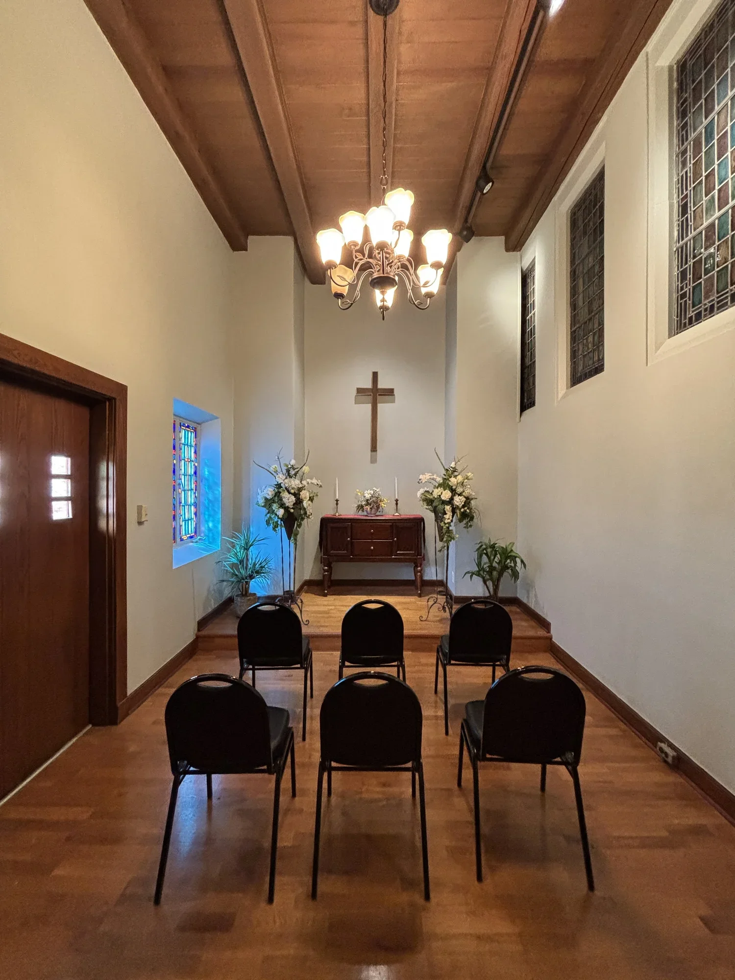 The interior of a small church with a wooden altar, decorated with flowers and candles, a cross on the wall behind, wooden floors, four black chairs in front, stained glass windows on the sides, and a chandelier hanging from the wooden ceiling.