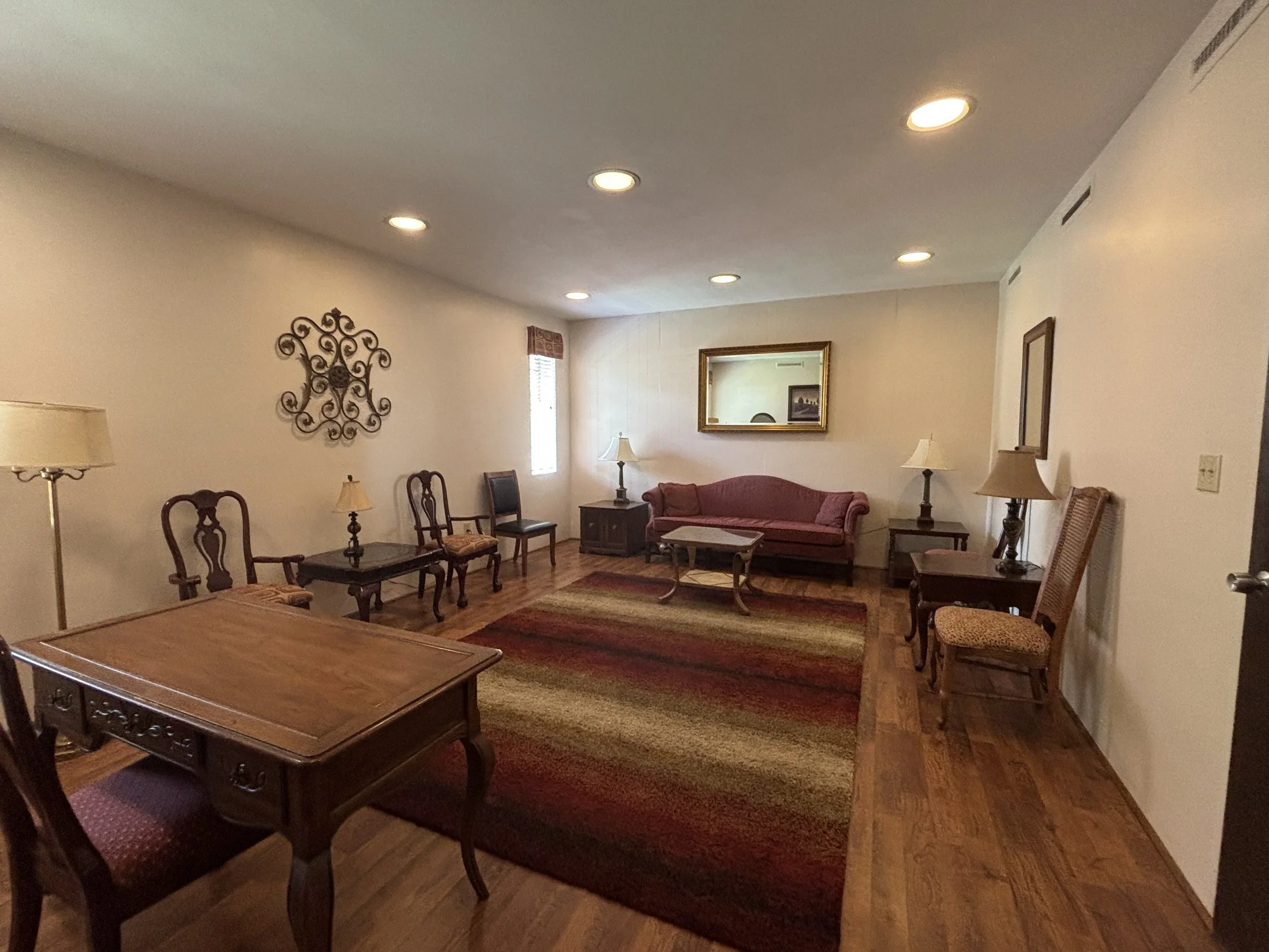 Living room with wooden furniture, including chairs and tables, a red upholstered sofa, lamps, a mirror, and wall decorations with beige walls and hardwood flooring.