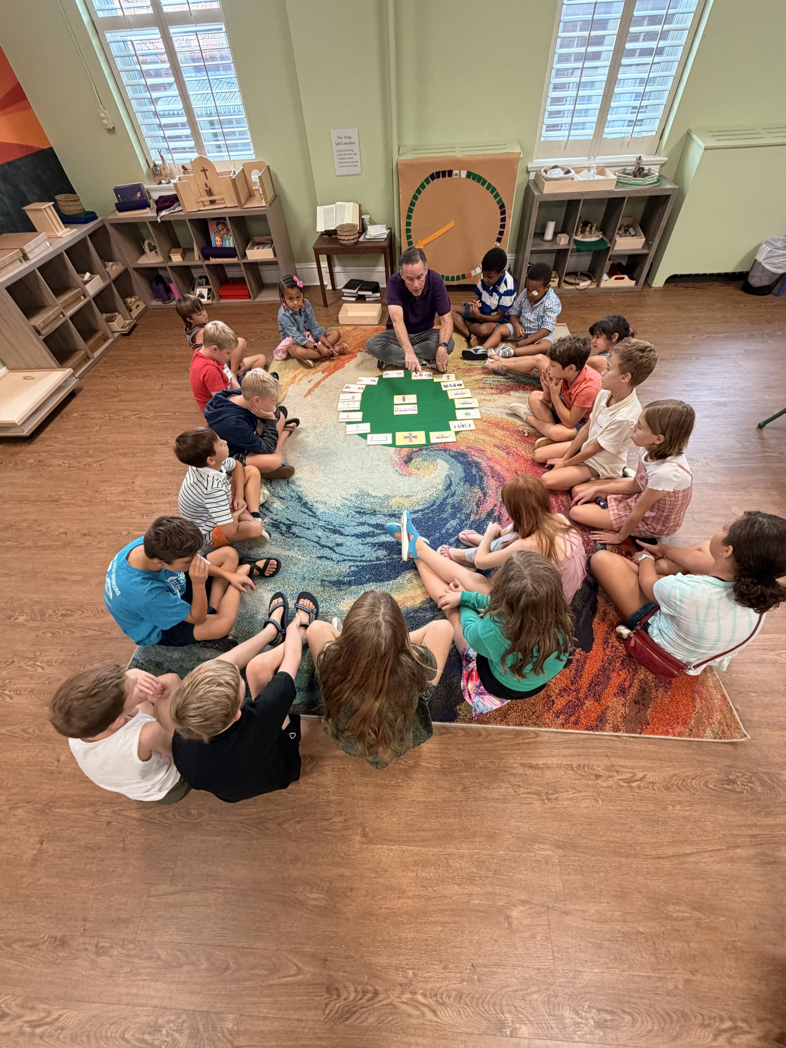 Children and an adult sitting on a rug in a circle in a classroom, engaging in a learning activity with cards. There are shelves with educational materials and windows with city views.