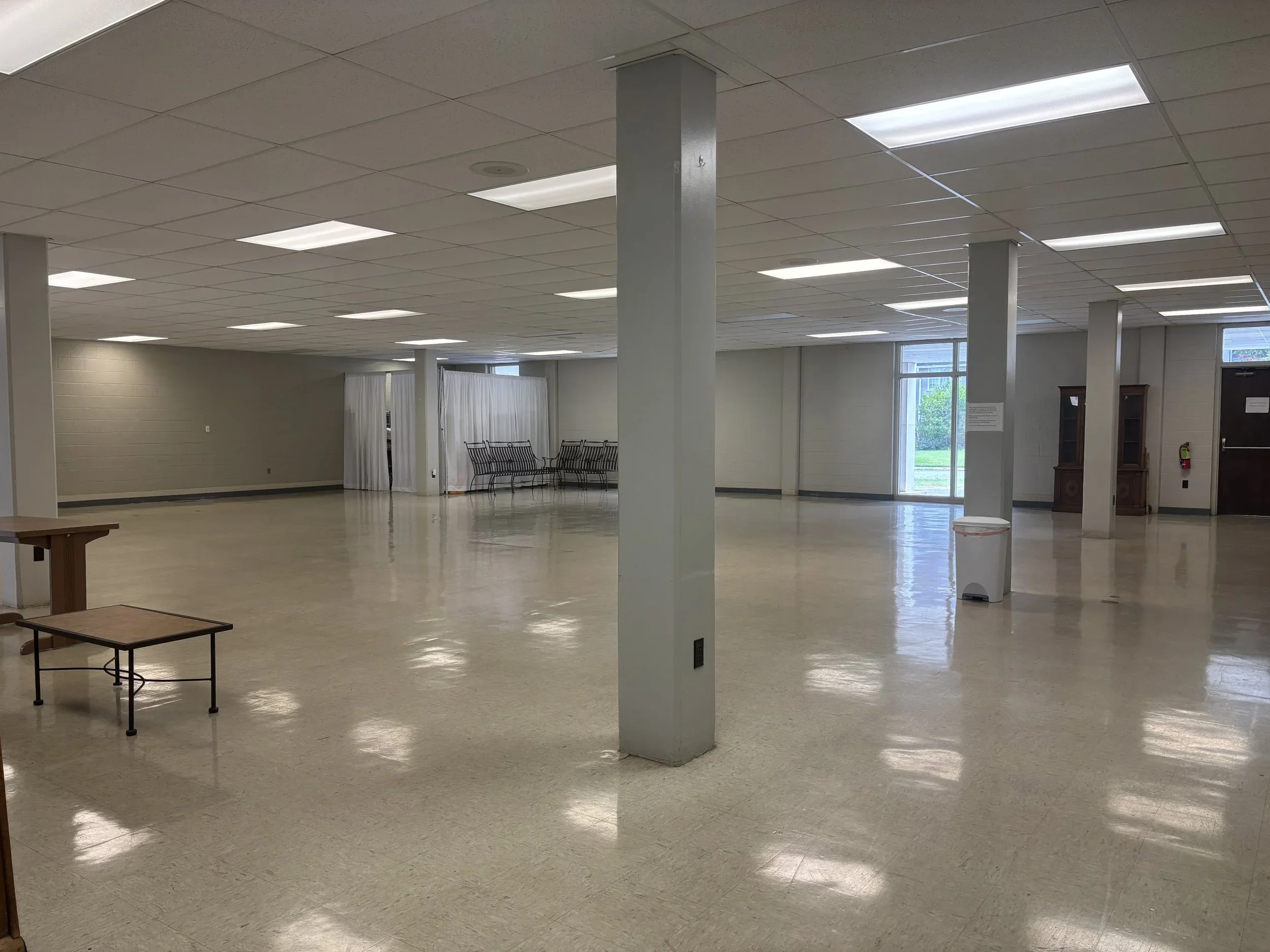 An empty indoor room with a tiled ceiling, fluorescent lighting, white walls, and large windows showing trees outside. There are benches, a small table, and a waste bin visible.