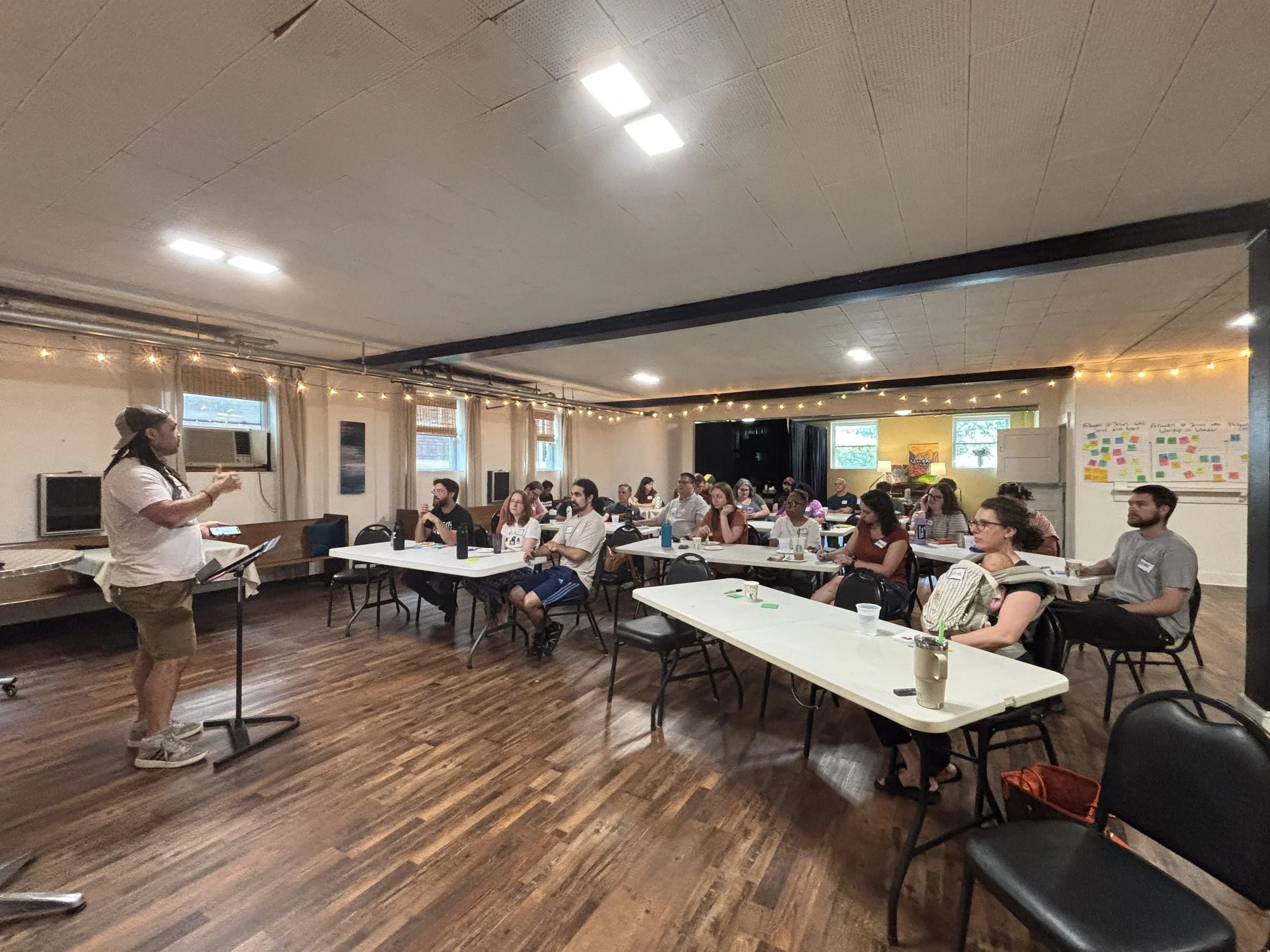 A group of people sitting at tables in a room with string lights and windows, listening to a person speaking at the front.