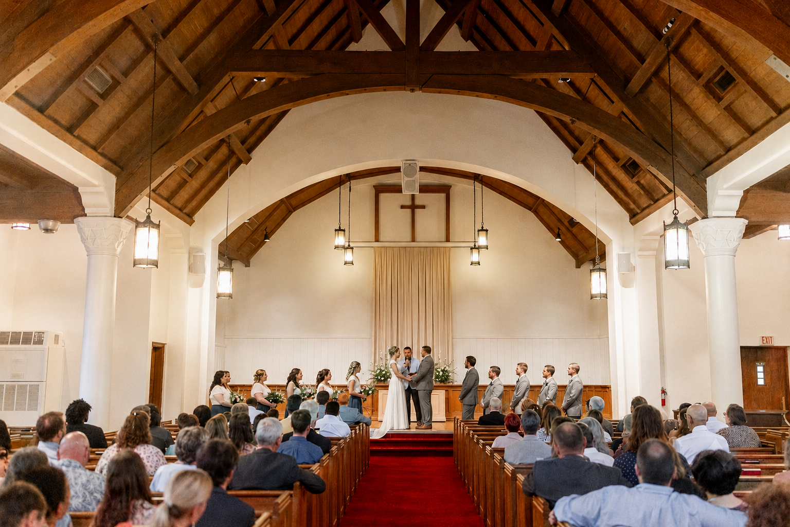 A wedding ceremony taking place inside a church with a wooden ceiling, white walls, and columns. The bride and groom are standing at the altar, exchanging vows. Bridesmaids and groomsmen are lined up on either side, and seated guests are watching the ceremony.