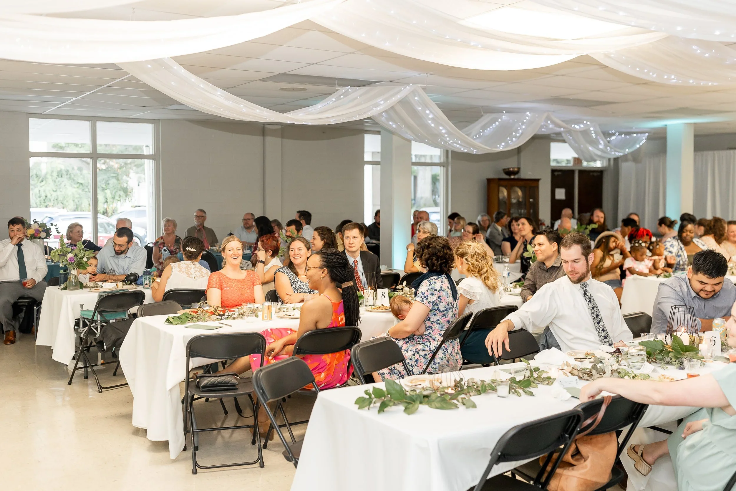 People attending a celebration or wedding reception sitting at decorated tables inside a hall with draped fabric and string lights on the ceiling.