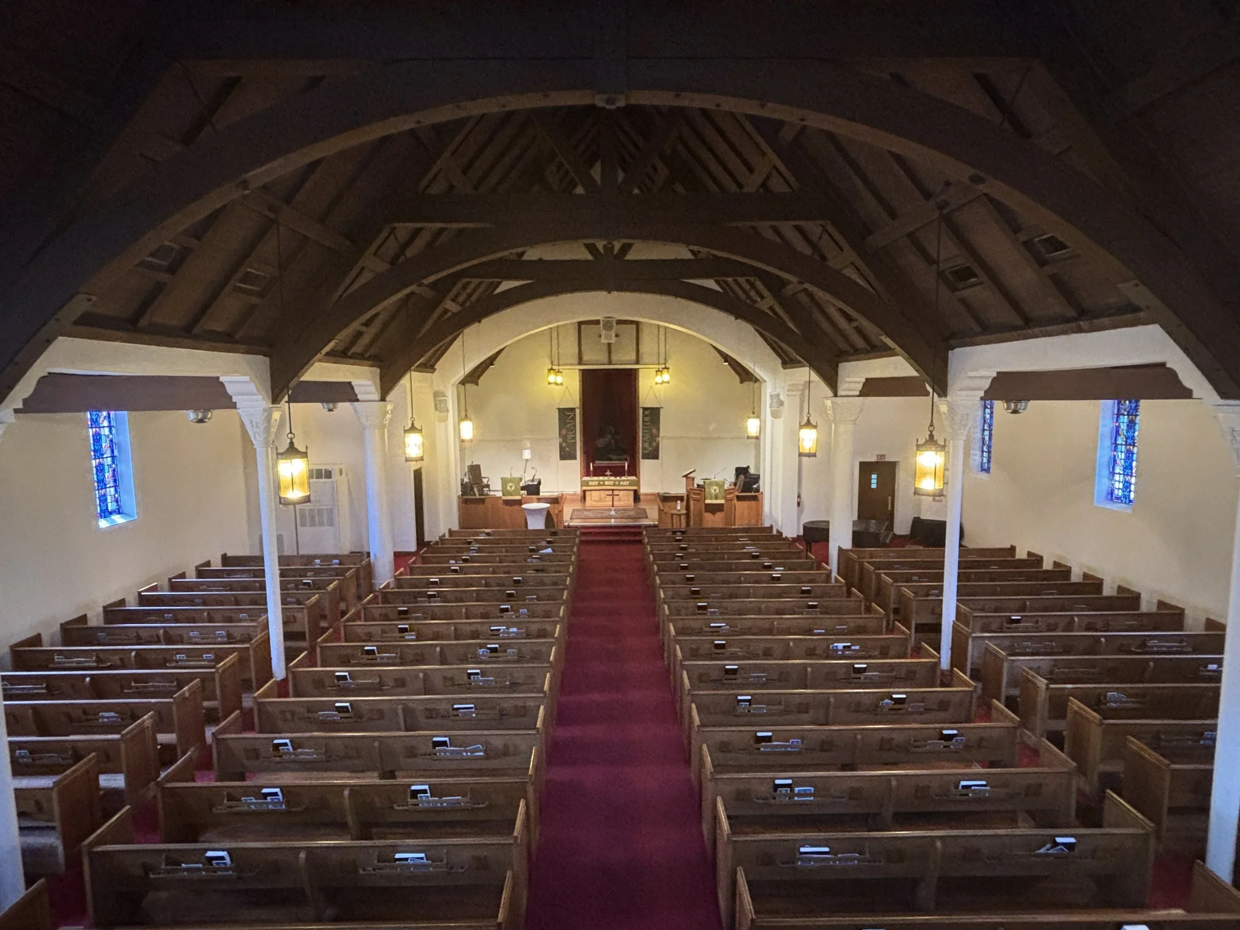 Interior of a church with wooden pews, a red carpet aisle leading to the altar, stained glass windows, and hanging lantern-style lights.