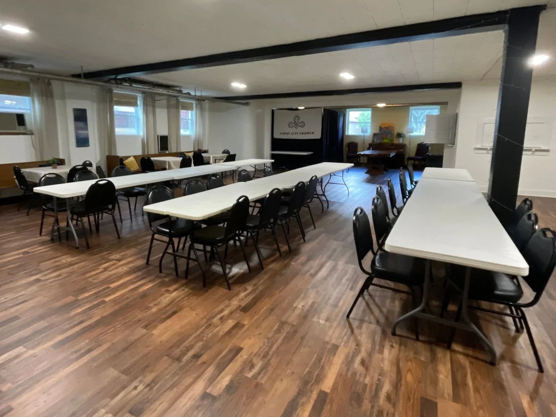 Empty meeting room with long white tables and black chairs, church banner in the background, and wood flooring.