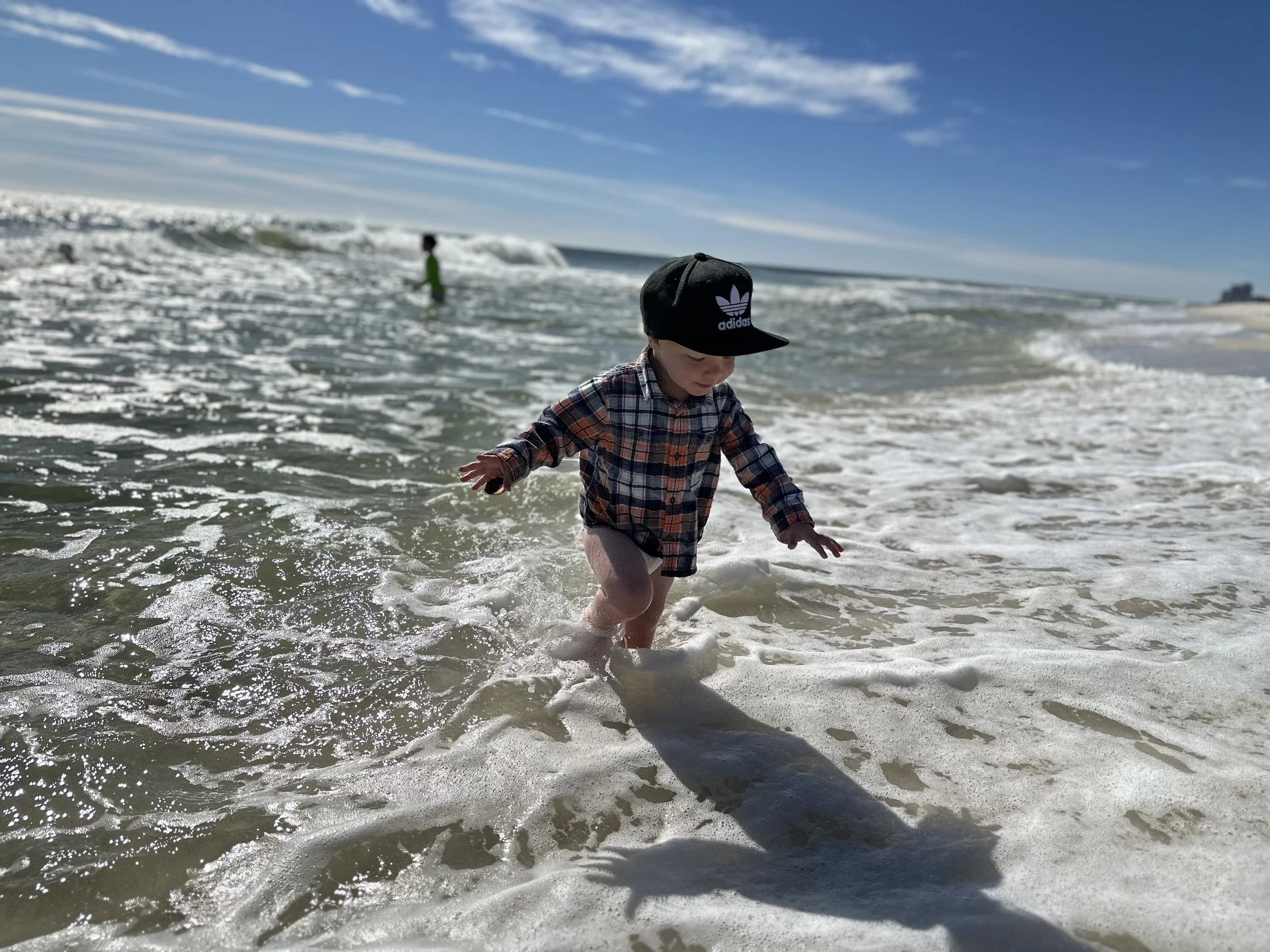A young child wearing a black Adidas cap and a plaid shirt playing in the shallow surf at the beach during daytime under a clear blue sky.