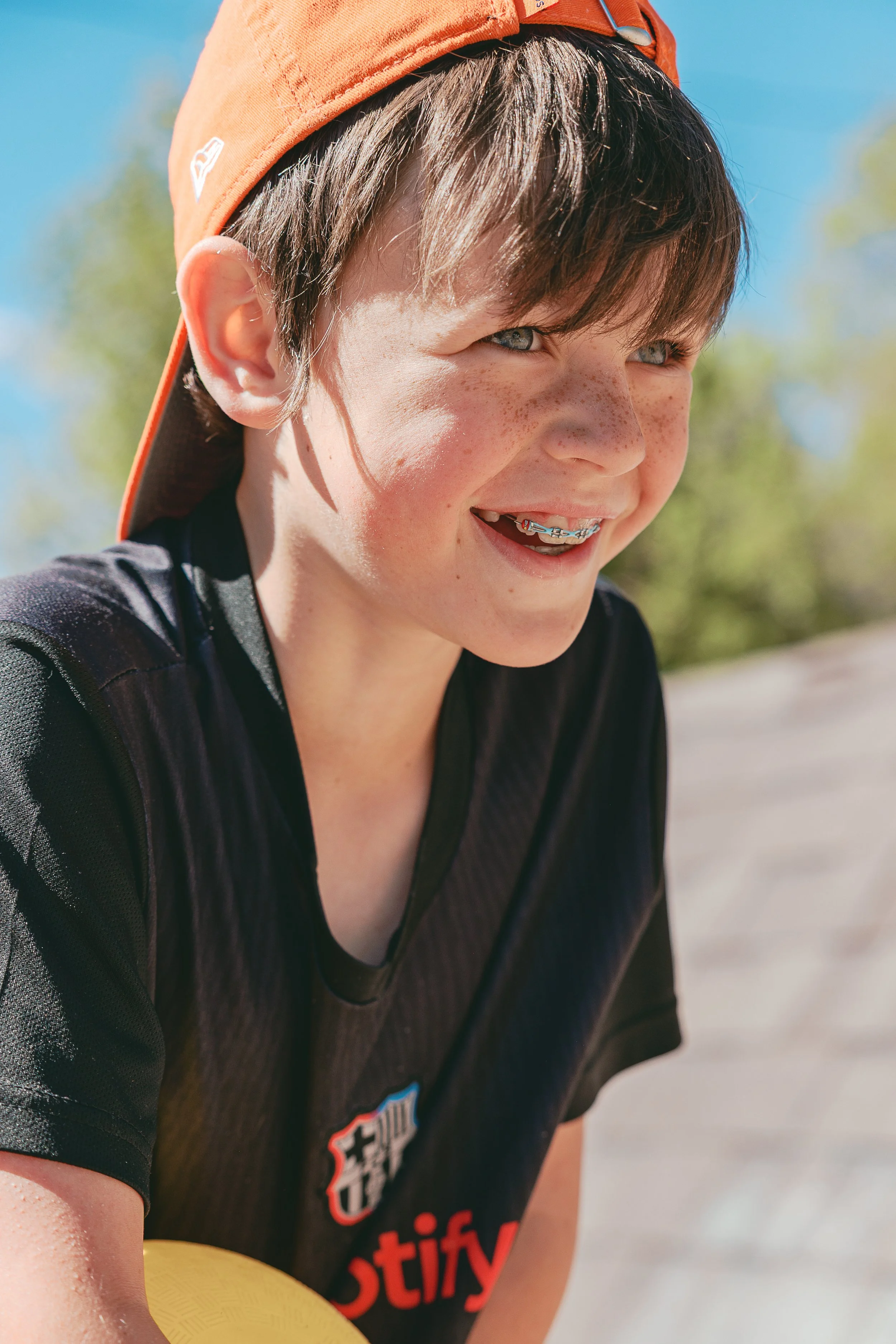 A young boy with blue eyes, freckles, and brown hair wearing an orange cap and a black sports shirt, smiling outdoors on a sunny day.
