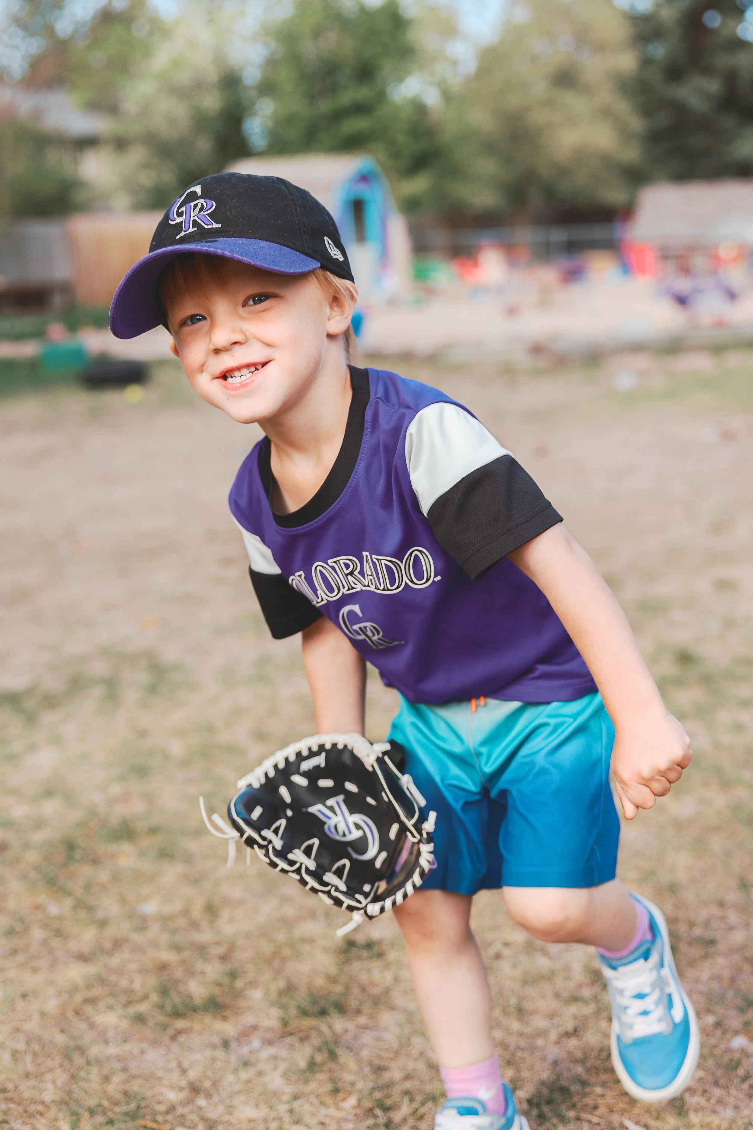 young boy playing baseball outdoors, wearing a black and purple Rockies cap, purple Rockies jersey, blue shorts, and blue sneakers with pink socks, holding a baseball glove