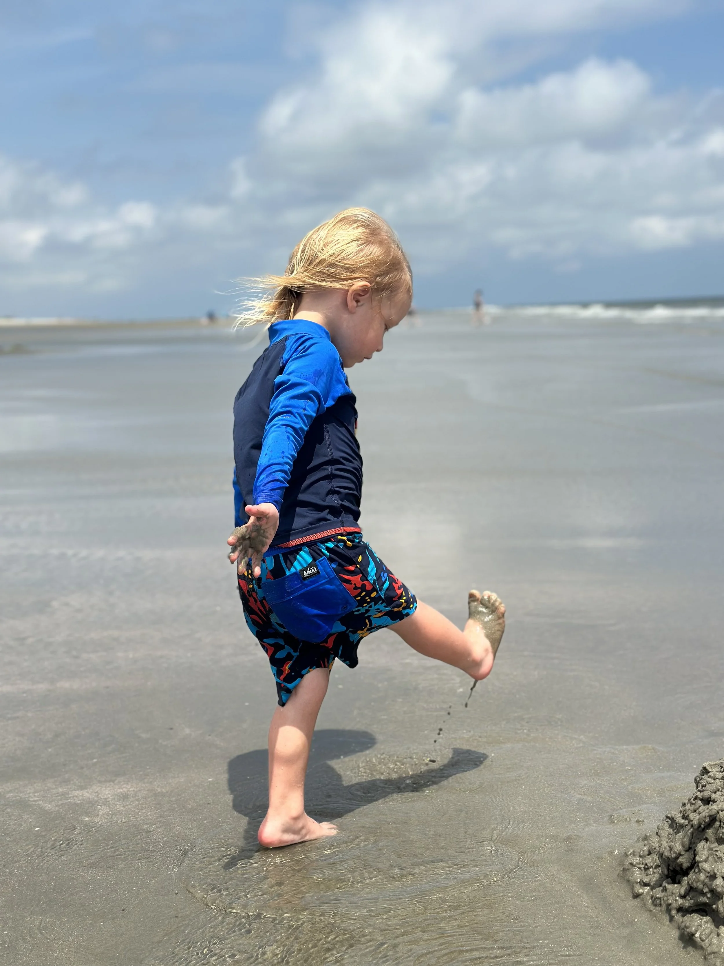 A young boy wearing a blue and black swimsuit playing with sand near the shoreline on a cloudy day at the beach.