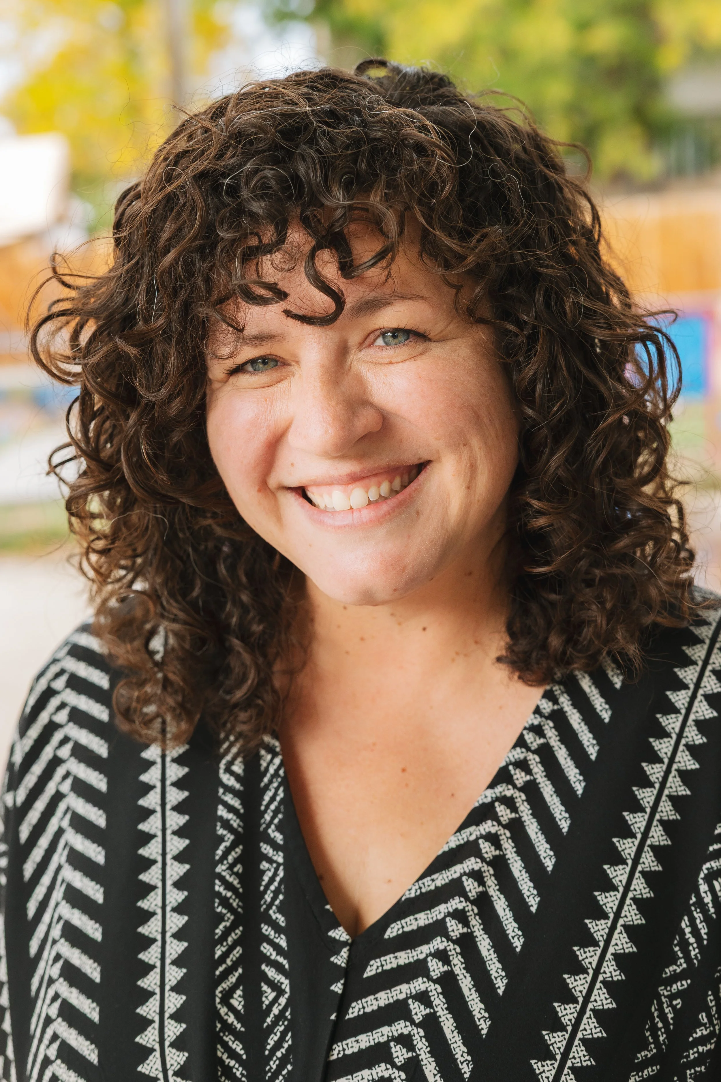 Close-up of a woman with curly brown hair smiling outdoors, wearing a black and white patterned top.