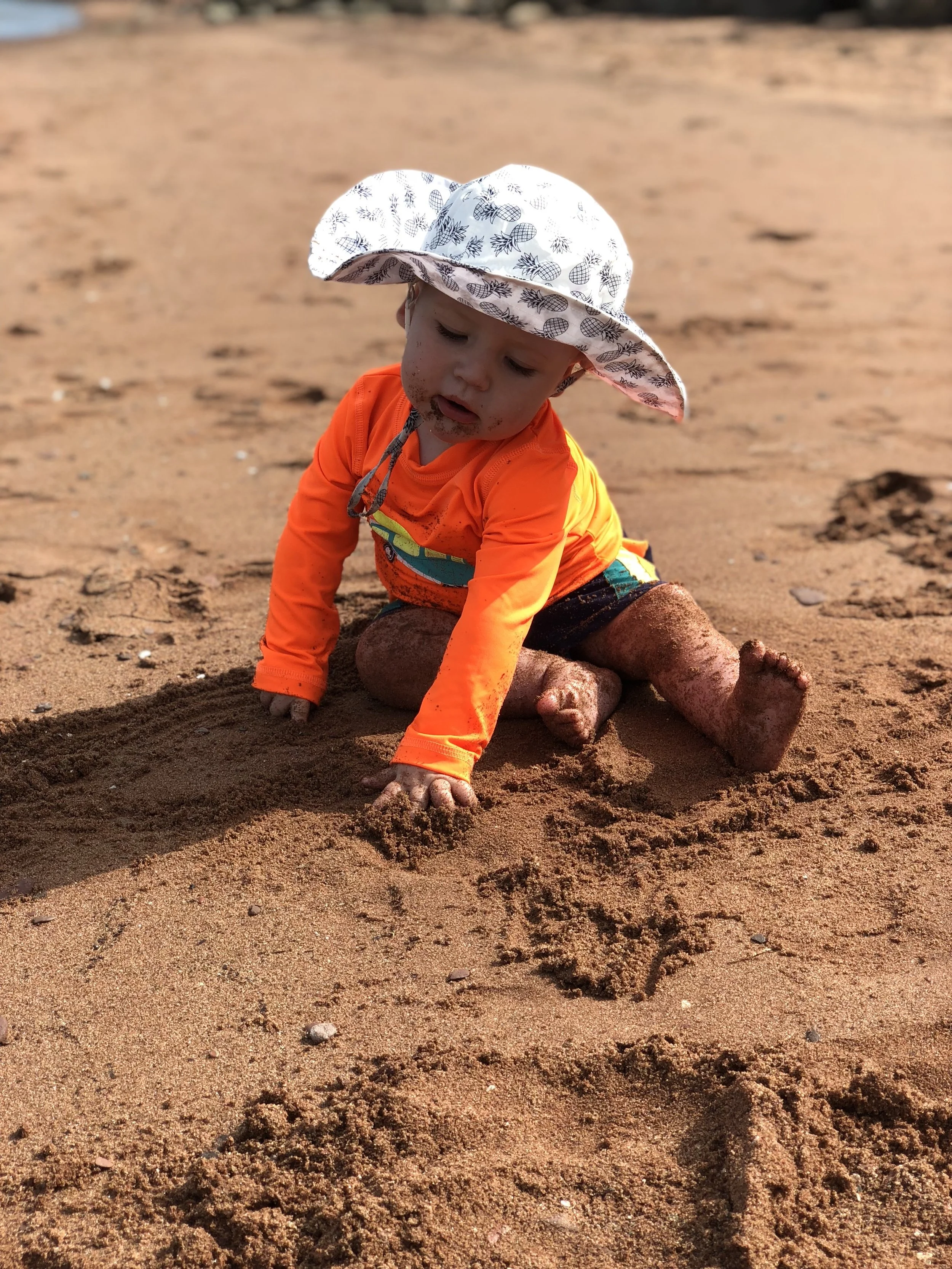 A young child in an orange shirt and patterned sun hat is playing on sandy beach, digging in the sand with their hands.