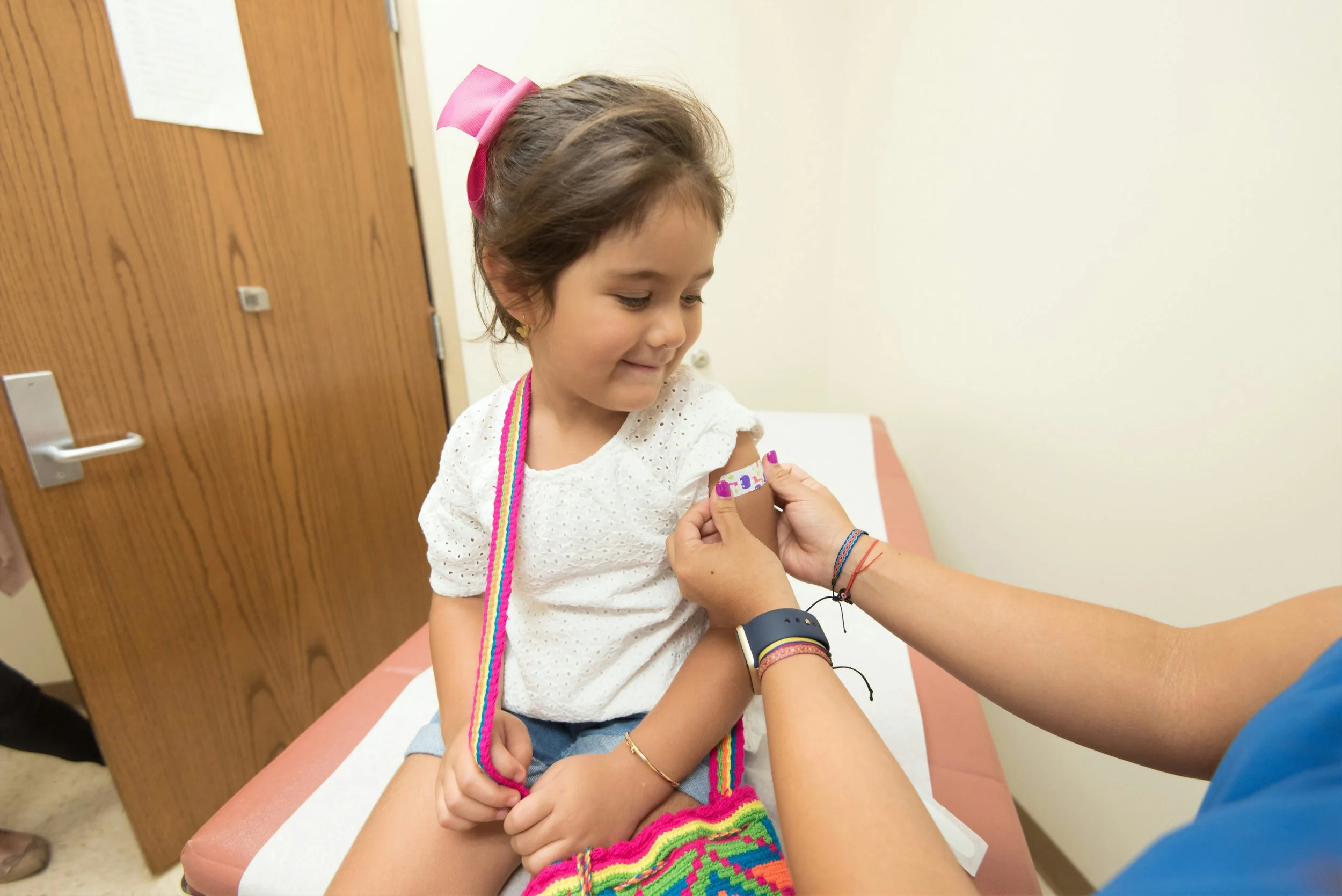 A young girl with short brown hair wearing a white top, rainbow-colored necklace, and rainbow socks, is sitting on a medical examination table, receiving a vaccination from a healthcare worker. The girl has a pink bow in her hair and is smiling while looking away. The healthcare worker's hand is administering the shot, with a bandage applied to the girl's arm. The setting appears to be a clinic with a wooden door in the background.