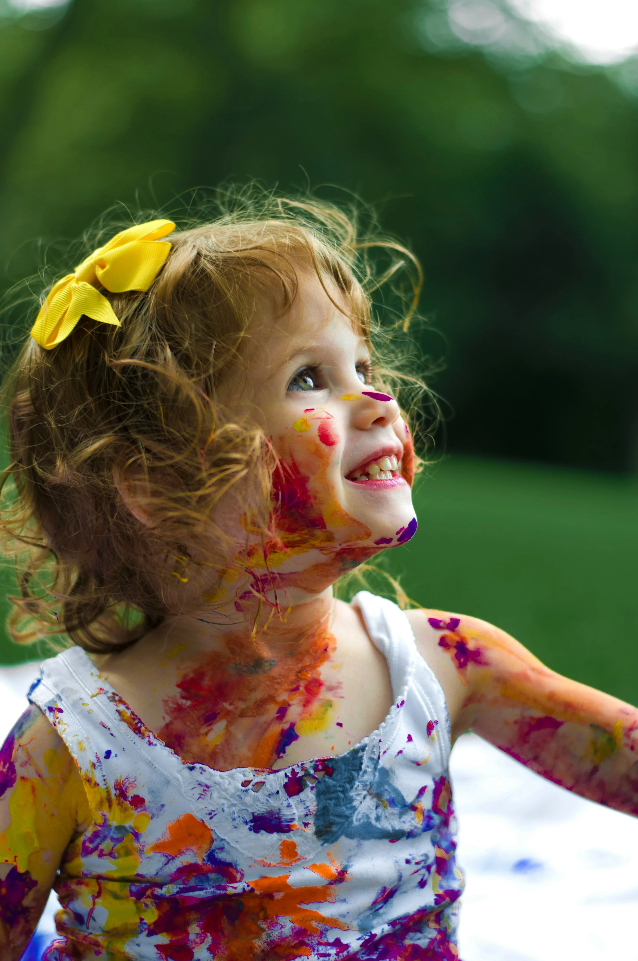 A young girl with curly red hair and a yellow bow in her hair is smiling outdoors. Her face and clothes are covered in colorful paint, and she appears to be enjoying a painting or messy play activity during a bright day.