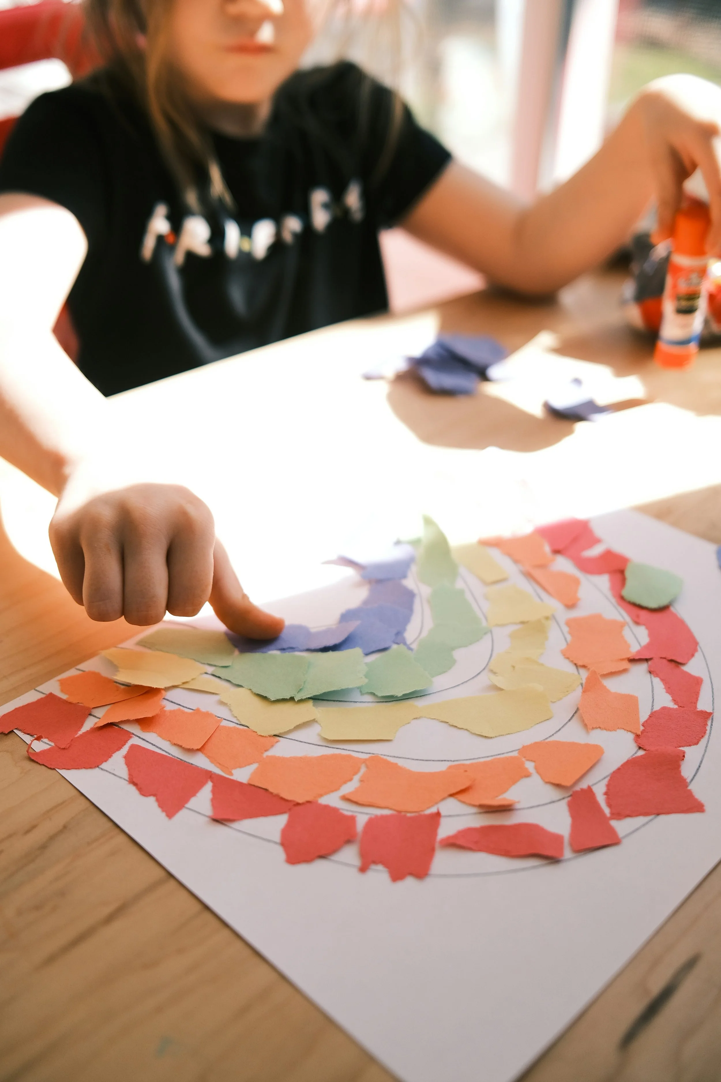Child creating a colorful autumn-themed collage with torn paper pieces on a table near a window.